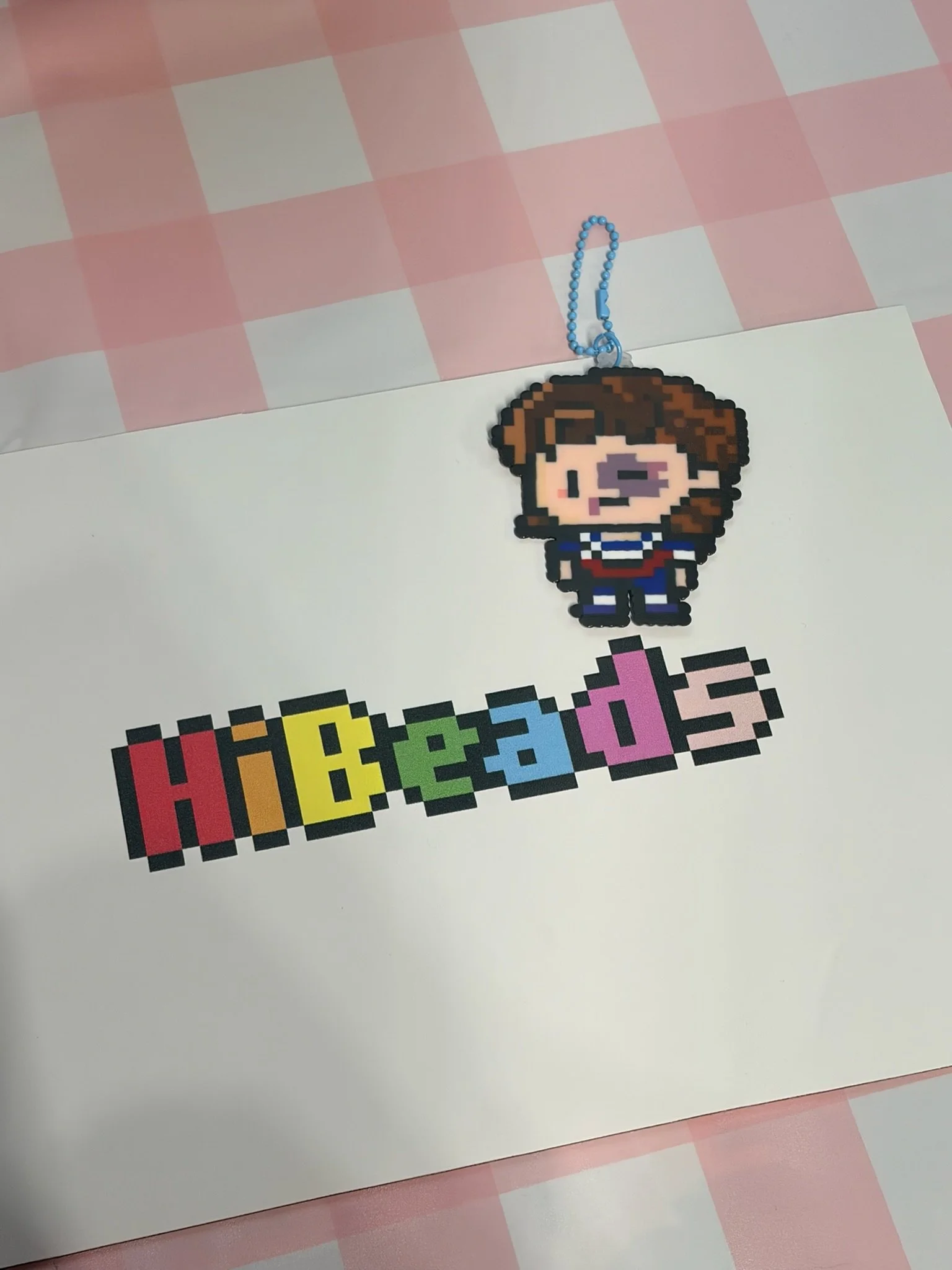 Pixel art keychain of a girl with brown hair, wearing a striped shirt, above the colorful word "Hobbies" on white paper, on a pink and white checkered tablecloth.