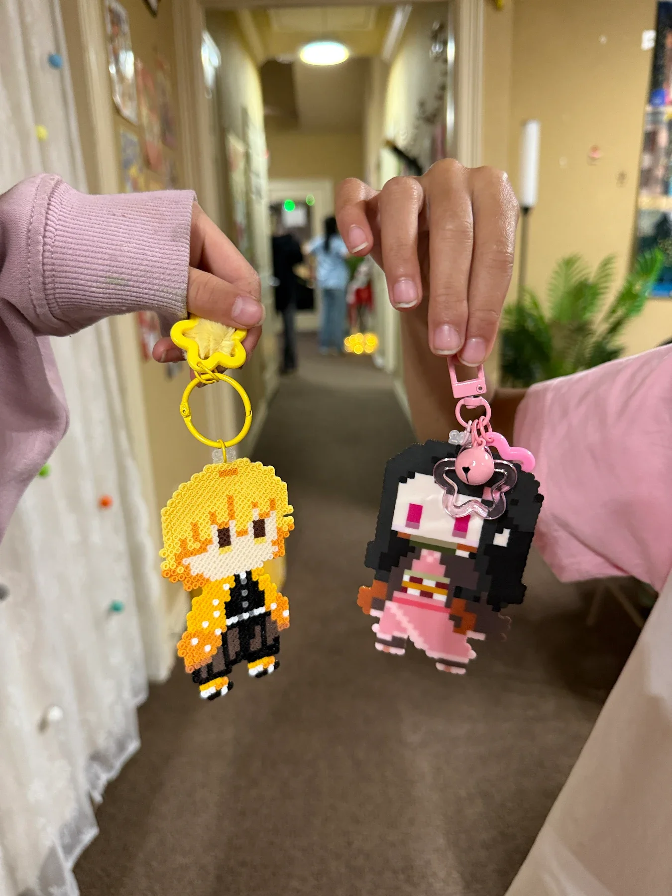 Two hands hold pixel art character keychains near a hallway. The left keychain is a character with orange hair, black outfit, and yellow accents, attached to a yellow star-shaped clip. The right keychain is a character with black hair, pink outfit, and pixelated features, attached to a pink clip.