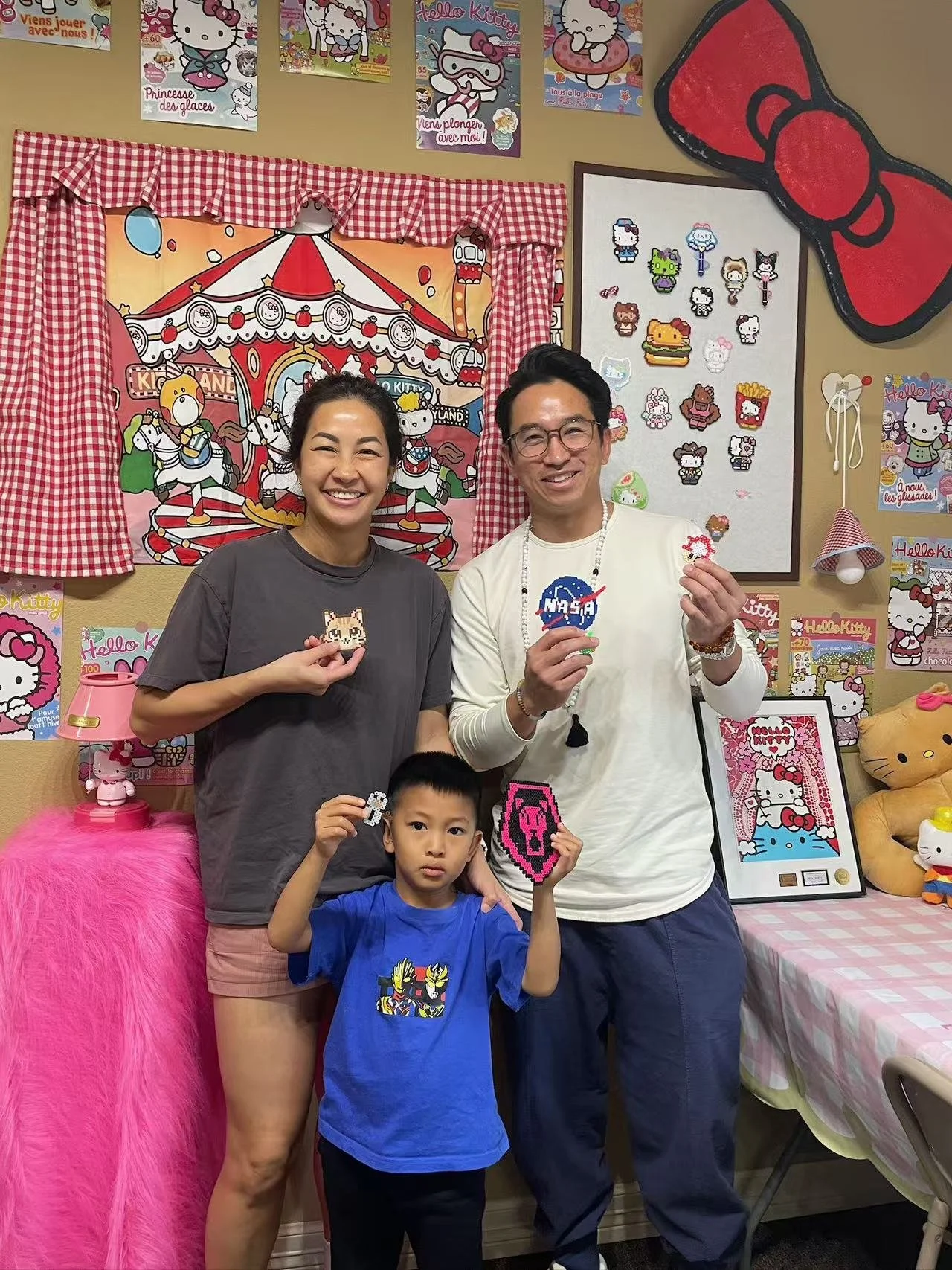 Three people, two adults and one child, smiling and holding Hello Kitty-themed pixel art crafts inside a decorated room with Hello Kitty posters, plush toys, and artwork on the walls.