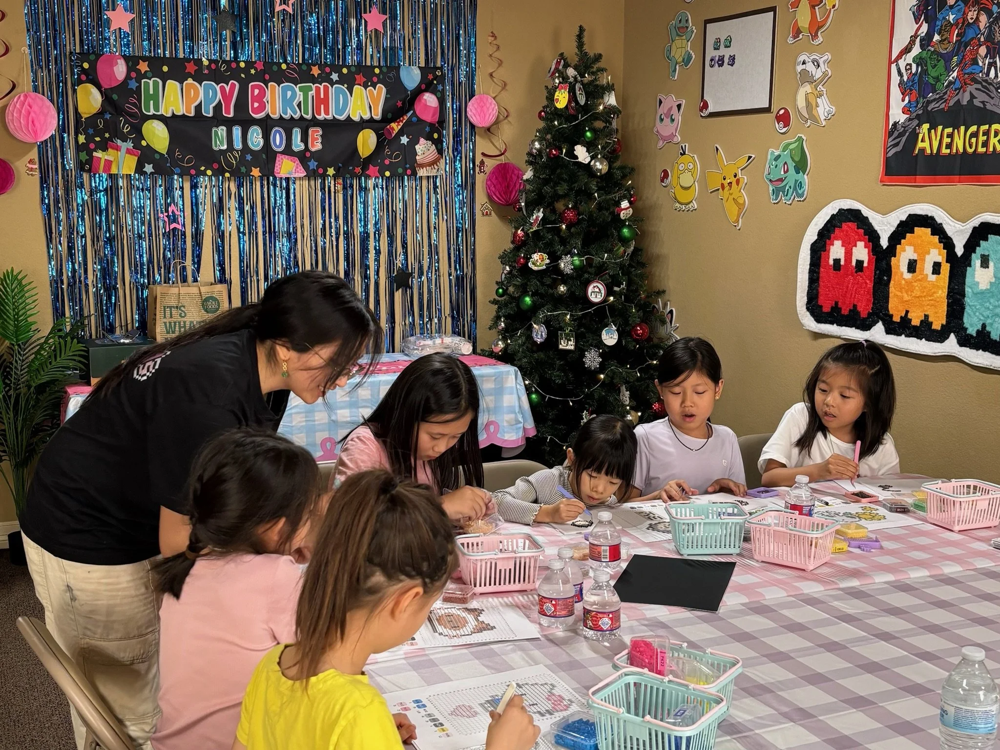 Children gathered around a table decorating for a birthday party, with a Christmas tree in the background and Pokémon themed decorations on the wall.