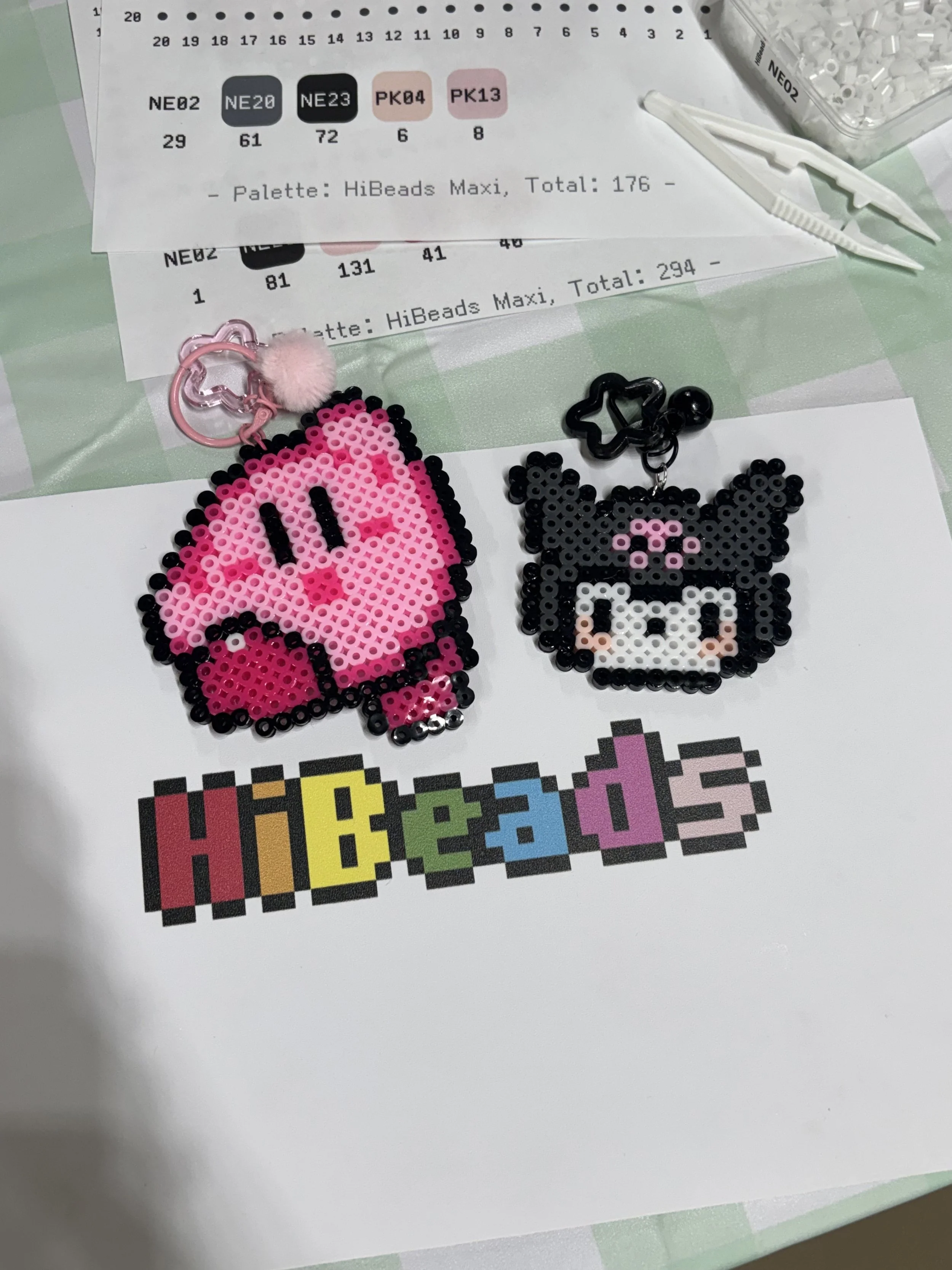 Colorful pixel art accessories, including a pink cute character keychain and a black and white animal face keychain, placed on a white paper with the word 'Hibeads' in multicolored pixel font, on a craft table.