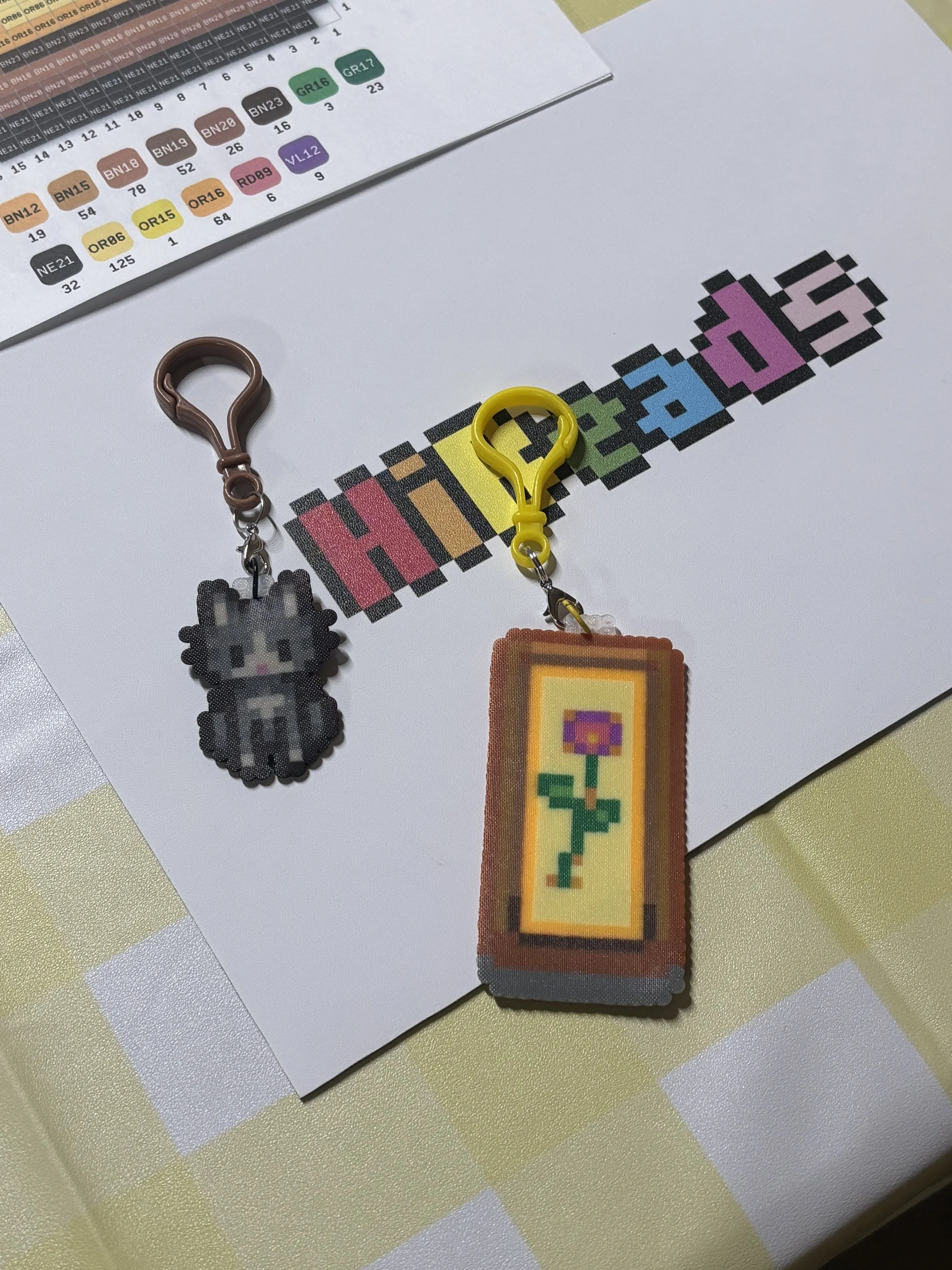Pixel art keychains of a cat and a flower placed on a white paper with colorful text that spells 'Hibeads'. A color chart with numbers and color codes is partially visible at the top of the image.