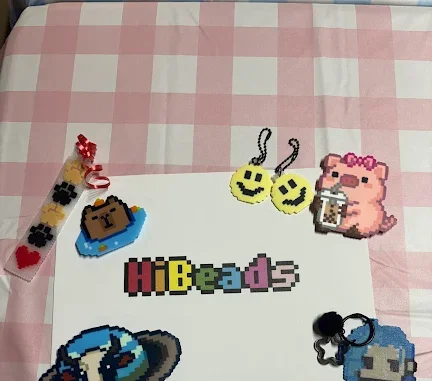 Stationery and pixel art accessories on a pink and white checkered tablecloth, including a HiBeads sign, pixelated emojis, and character charms.