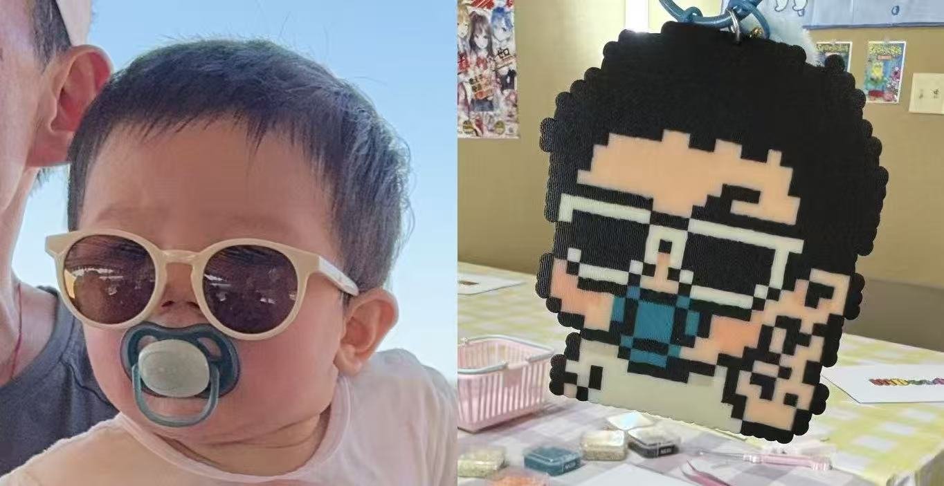 Side view of a young child with sunglasses and a pacifier, held up by an adult. Next to them is a pixel-art style emoji decoration with black, white, and blue colors, featuring glasses and a blue shirt, placed on a table with small containers and craft supplies in the background.
