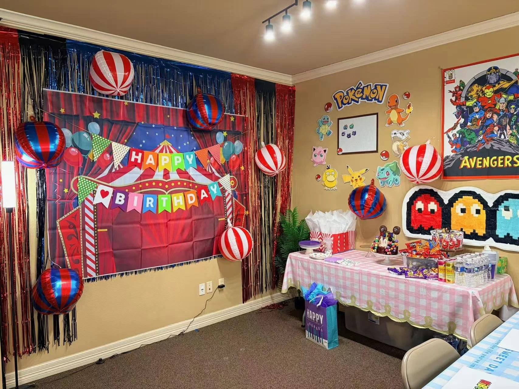 Decorated birthday party area with a carnival theme, featuring a red and blue backdrop with gold stars, multicolored flags and a banner that says 'Happy Birthday'. Red and blue striped hanging lanterns, assorted Pokémon and Marvel Avengers posters on the wall, and a table with snacks and gifts.