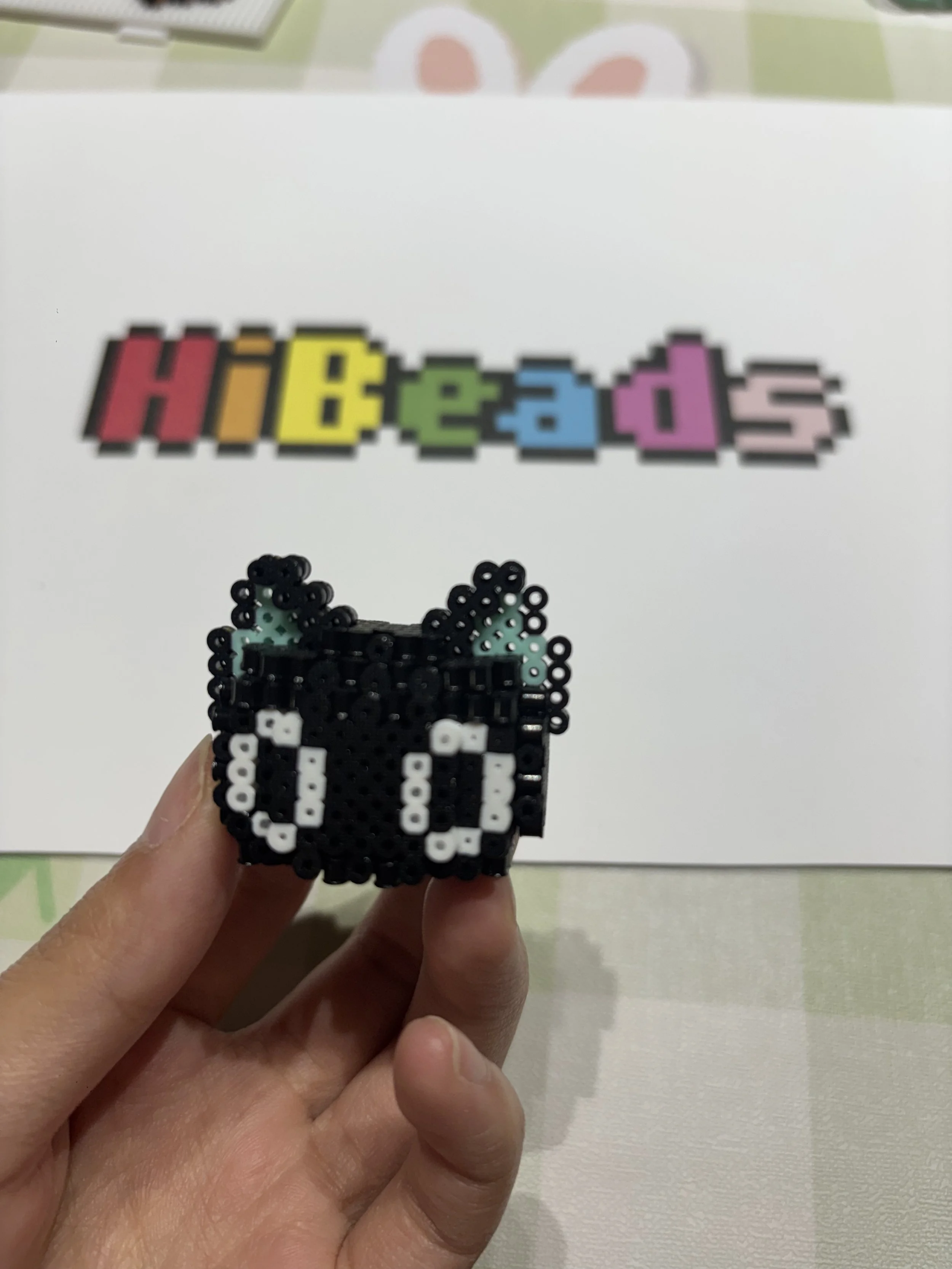 Person holding a pixel art bead creation of a black cat with teal ears and white eyes. In the background, a colorful pixelated word "HIDEADS" is on a white sheet of paper on a table with a pastel-colored tablecloth.