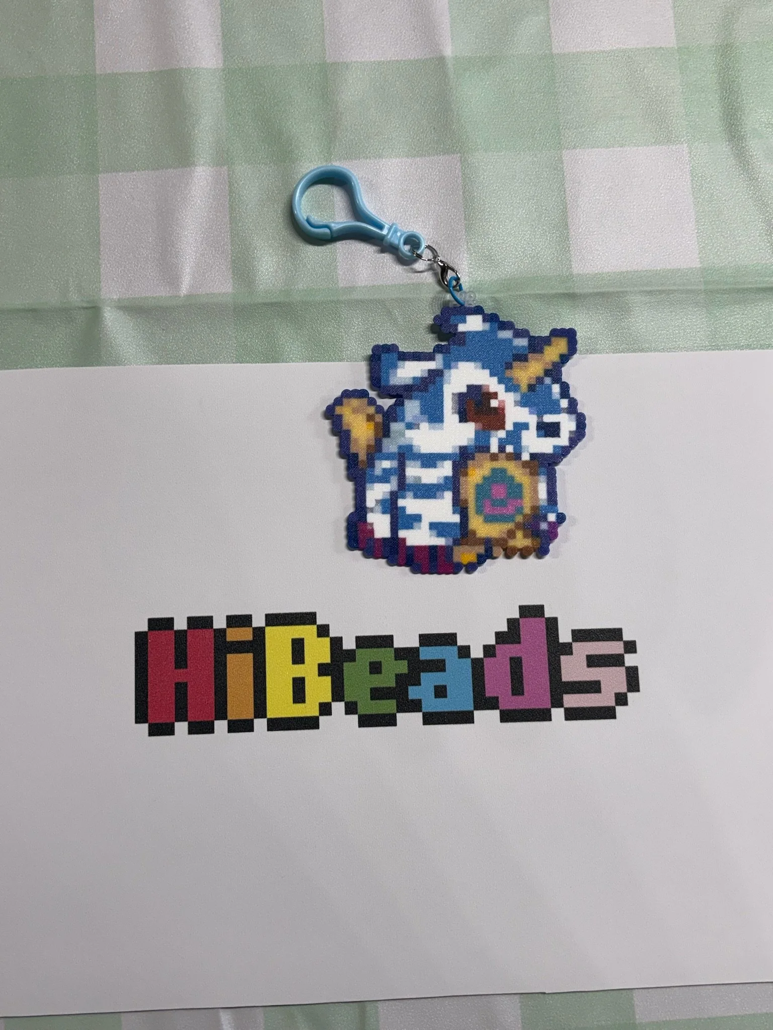 A pixel art keychain of a blue and white striped unicorn with a rainbow-colored heart on its chest, attached to a blue clip, placed above a sheet of paper with the word 'hobbies' in colorful pixelated letters.