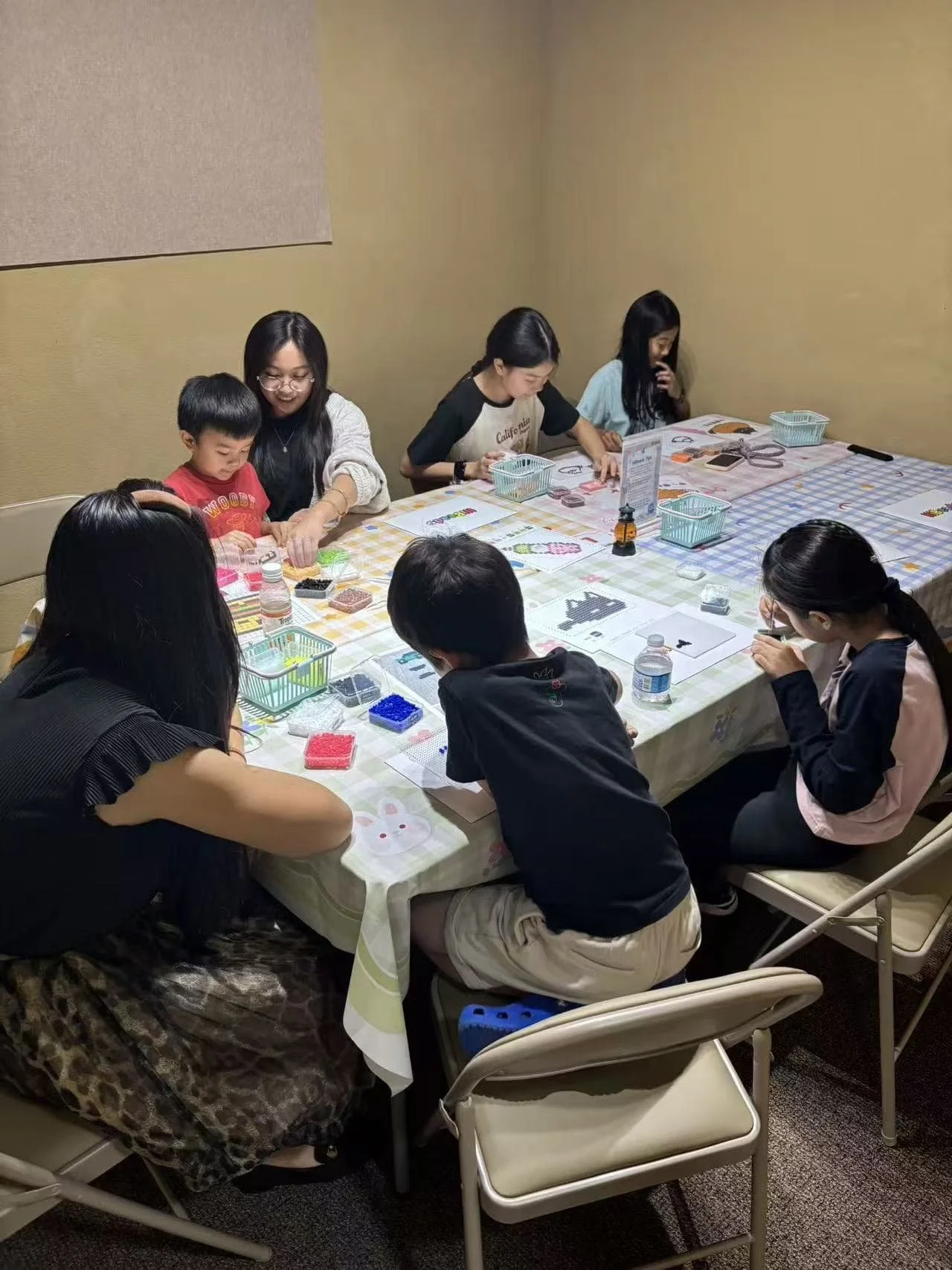 A group of children and adults sitting around a table engaged in arts and crafts, with supplies such as beads, paper, and tools on the table.