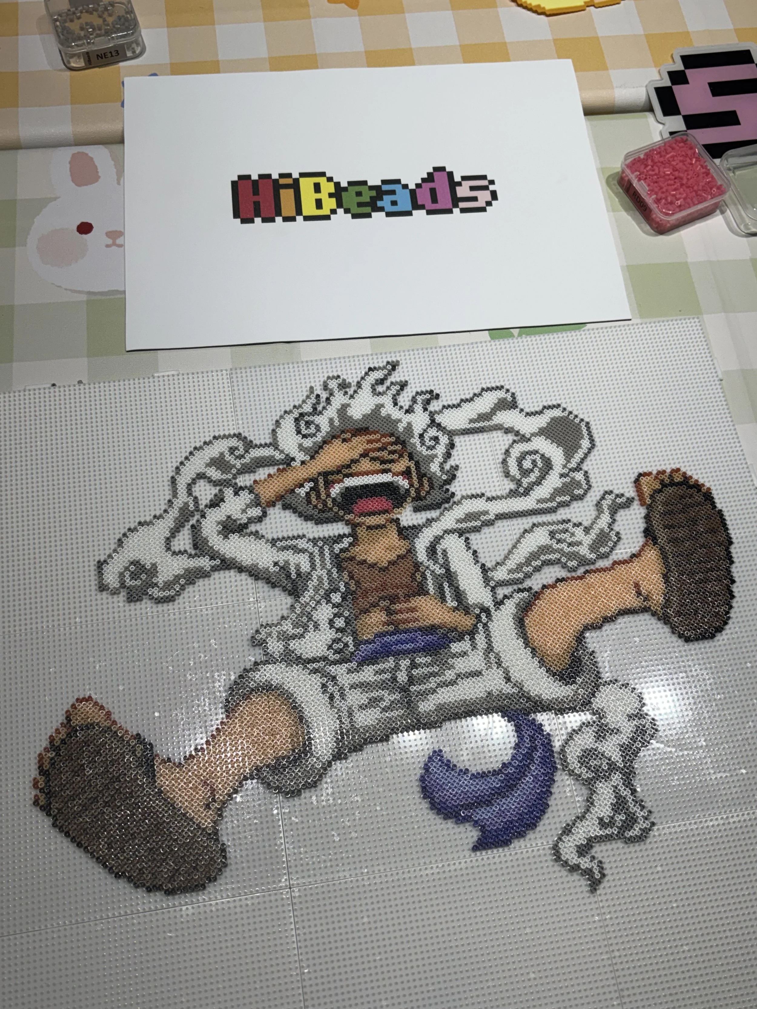 A pixel art mosaic of a surprised or shocked cartoon character, made with small circular beads placed on a pegboard. The character has gray hair, a tan shirt, shorts, and large shoes, and is sitting with one hand on his forehead and mouth open.