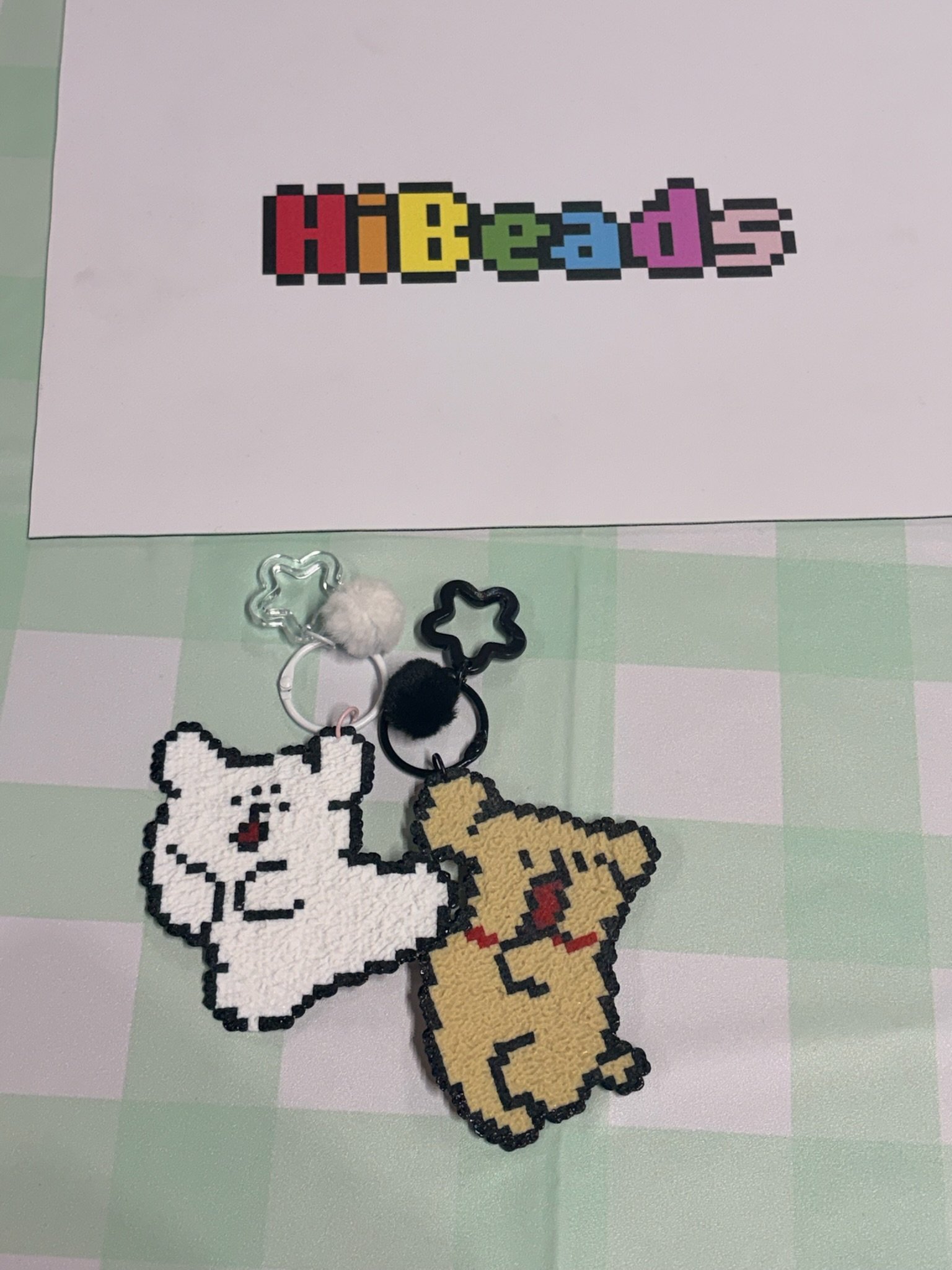 Pixel art keychains of white and yellow dogs, one with a red collar, on a light green and white checkered surface, with a Hibeads pixel art logo in the background.