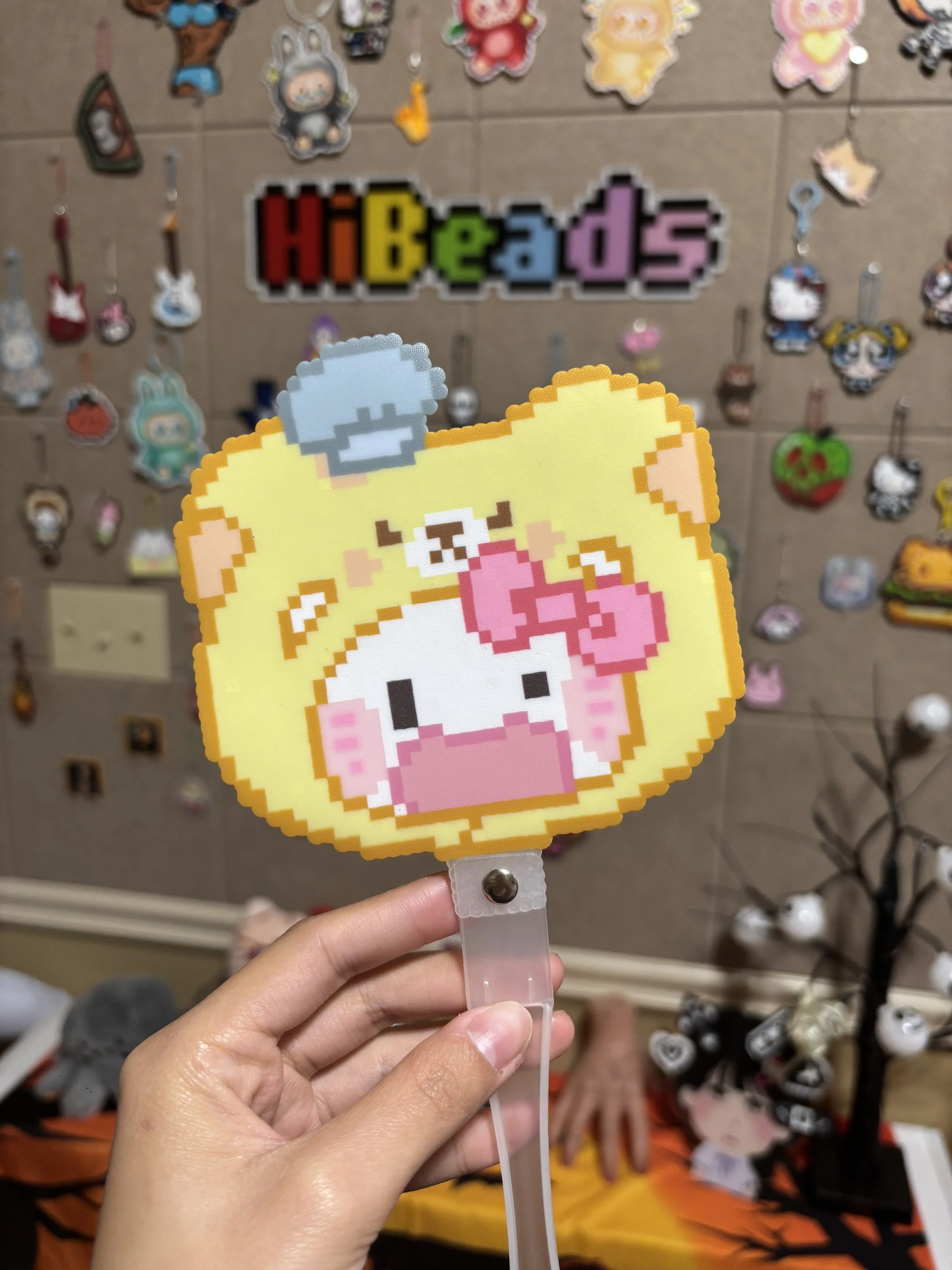Pixel art-style yellow pig with pink bow and piglet on its head, held by a hand in front of a wall decorated with various pixel art badges and a colorful 'HiBeads' sign.