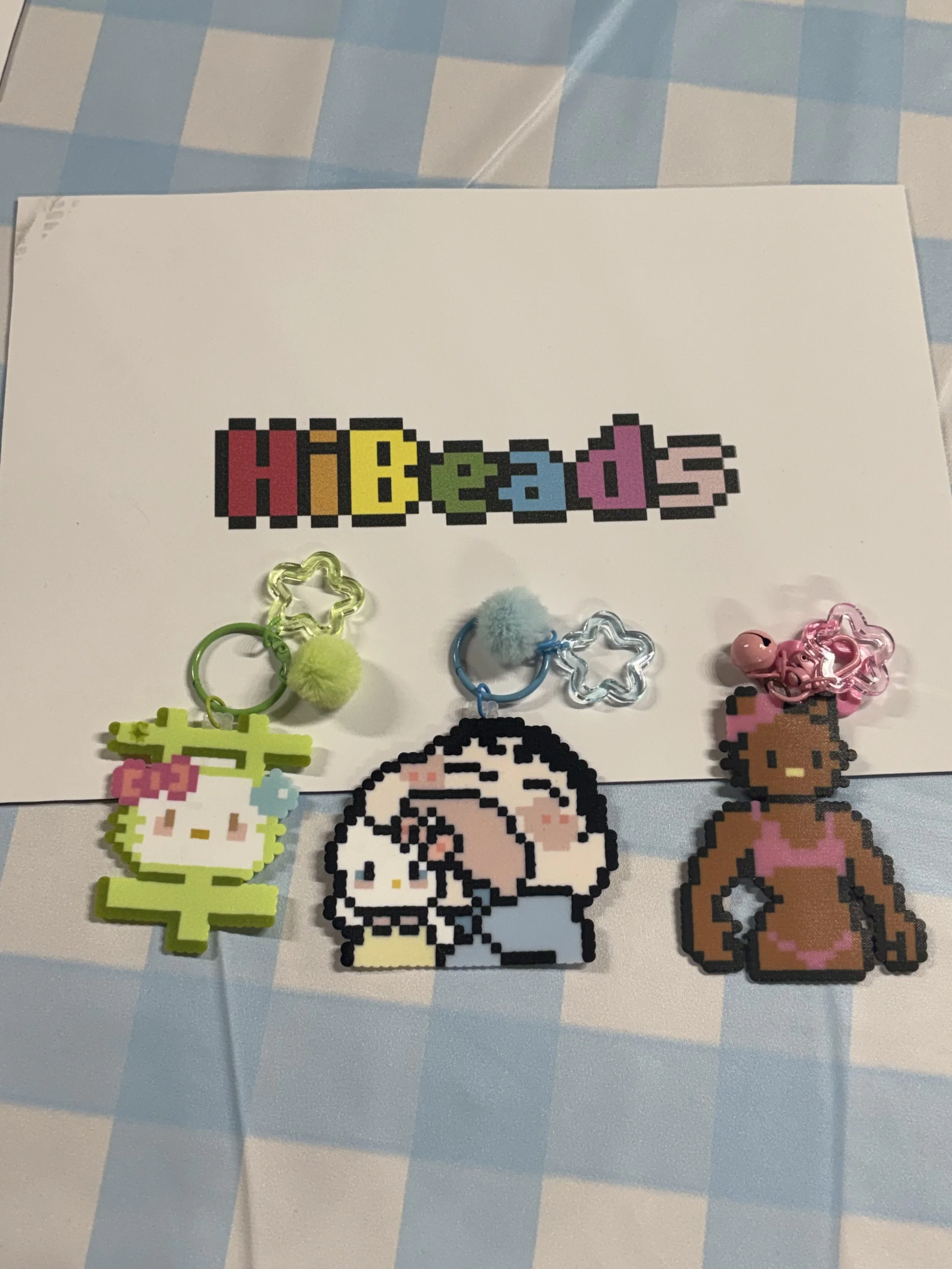 Colorful pixel art keychains with cartoon characters on a table. One keychain features Hello Kitty in a green outfit, another shows a girl with black hair and yellow accessories, and the third depicts a woman with dark skin and pink clothing.