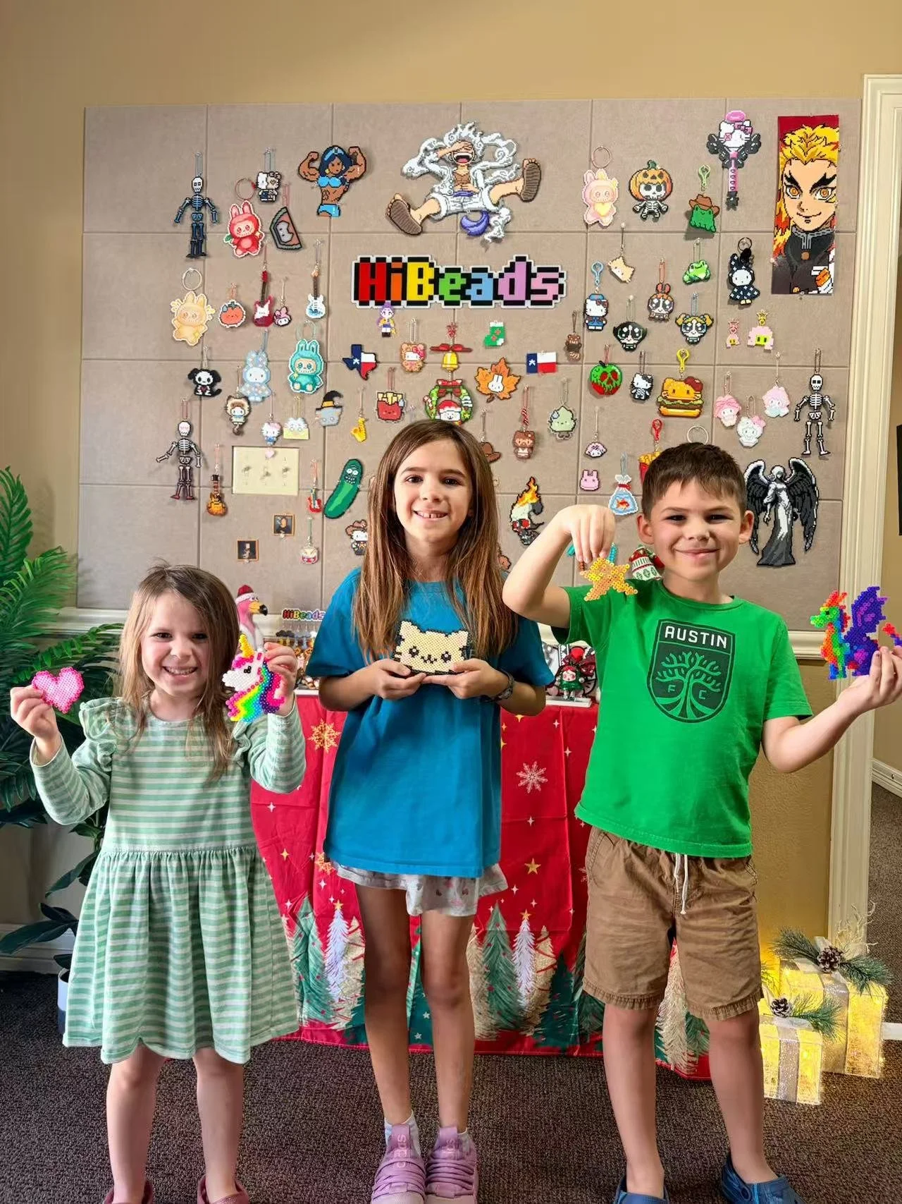 Three children standing in front of a display of pixel art beaded keychains and ornaments, holding some of their handmade beads, with a red holiday-themed tablecloth and Christmas decorations.