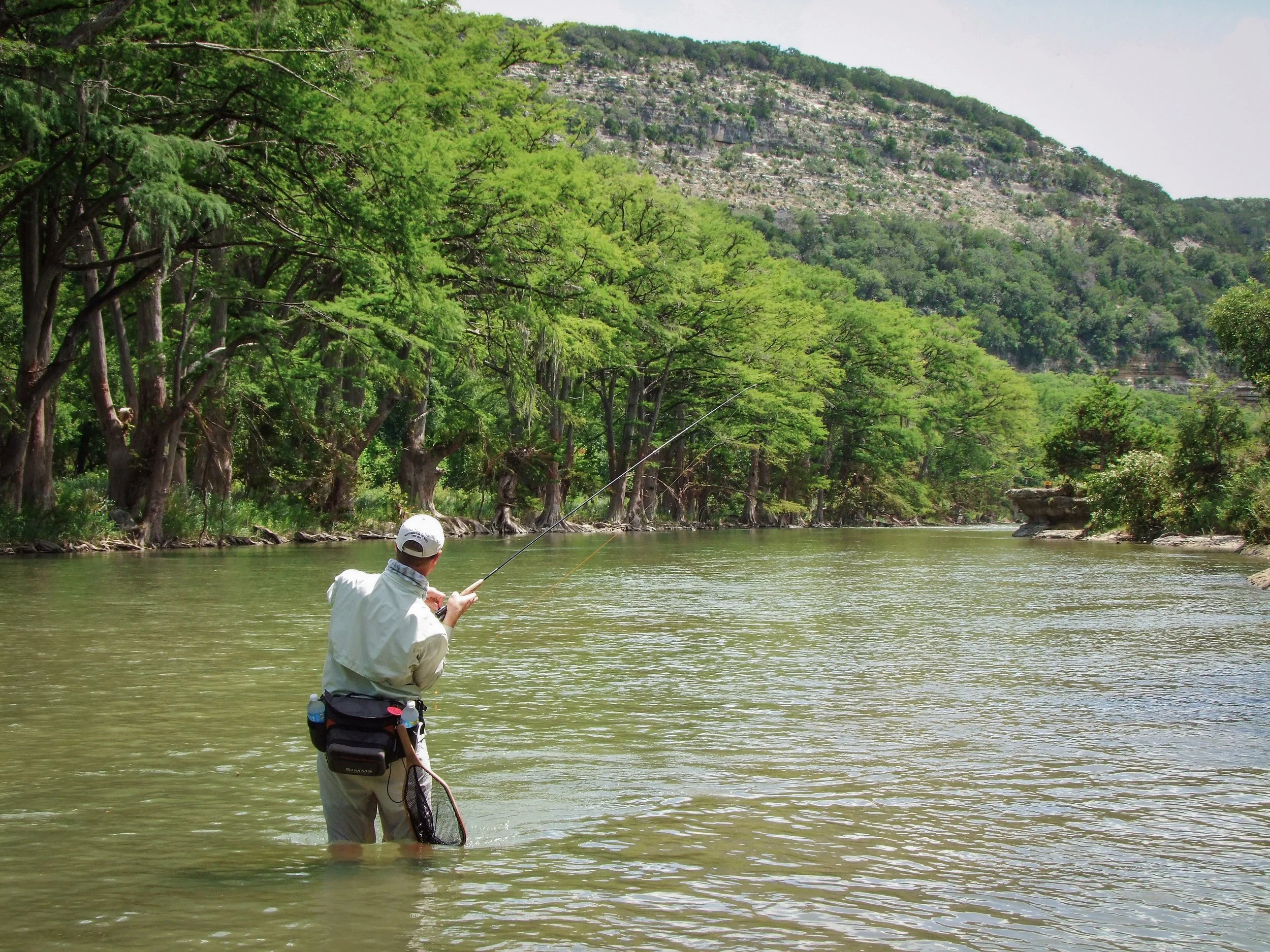 A person in fishing gear standing in a river, casting a fishing line with trees and a hillside in the background.