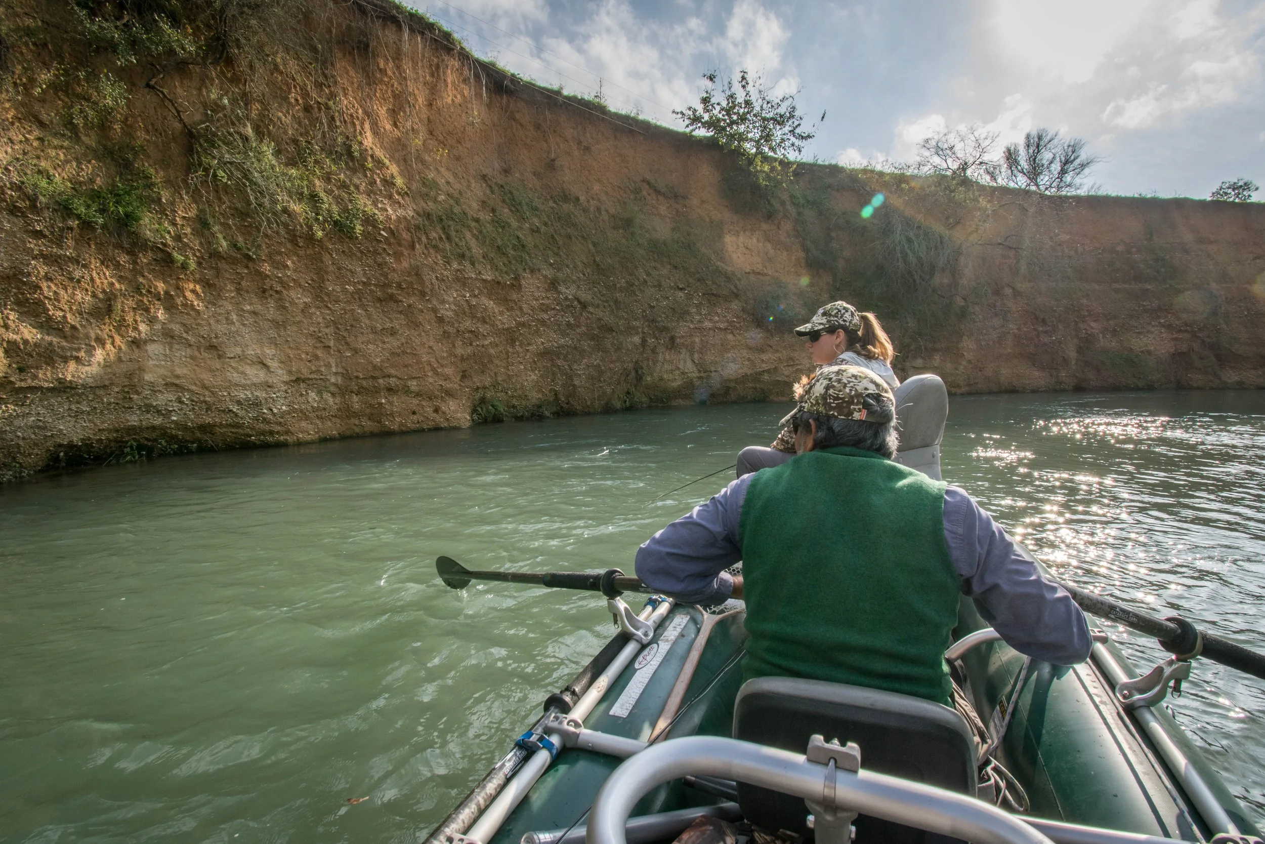 Two people in camouflage hats and sunglasses fishing from a boat on a river with rocky cliffs and trees in the background