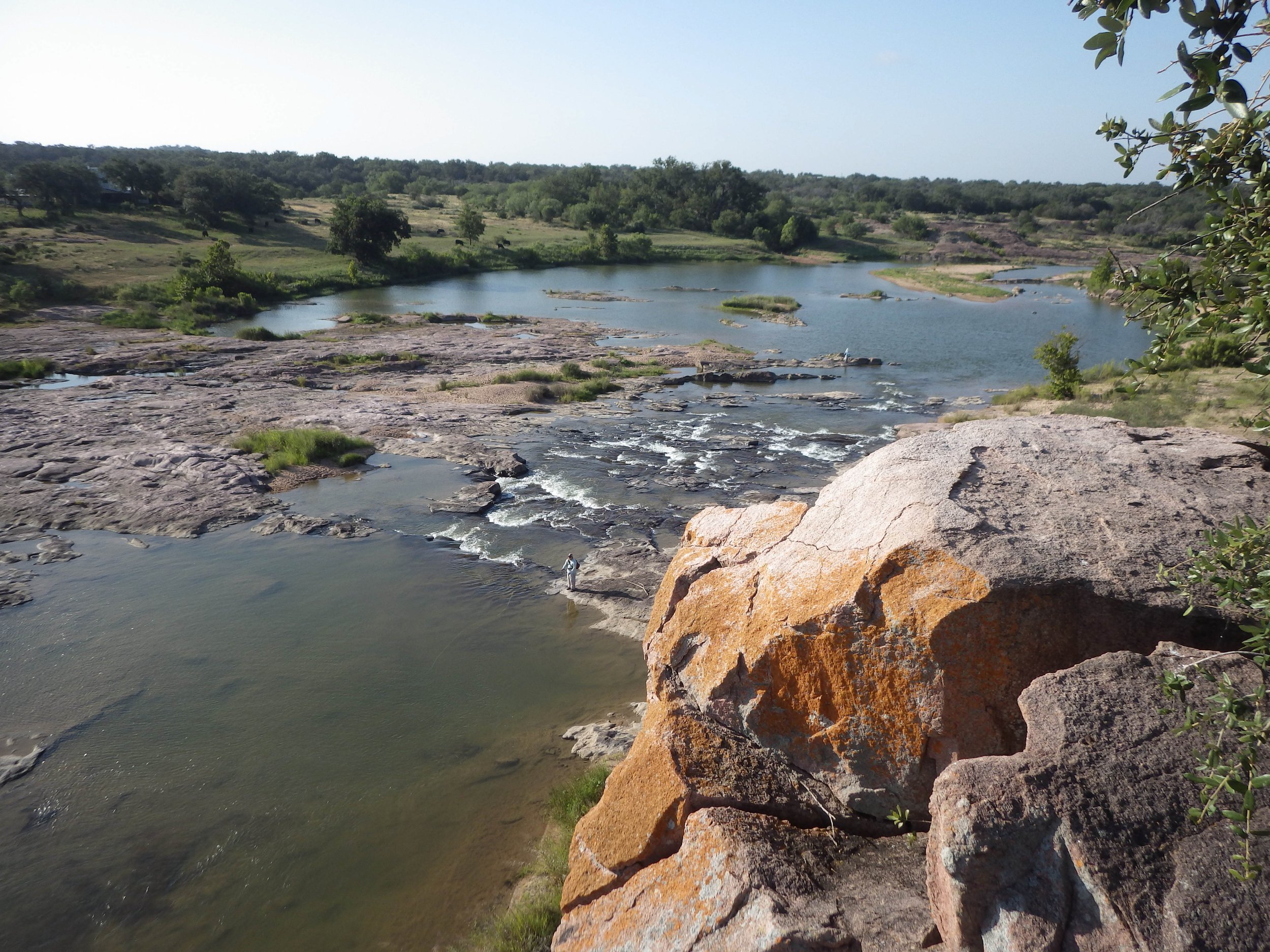 View of a river flowing over rocks with a lone fisher standing on a rock in the water, surrounded by green trees and hills under a clear sky.