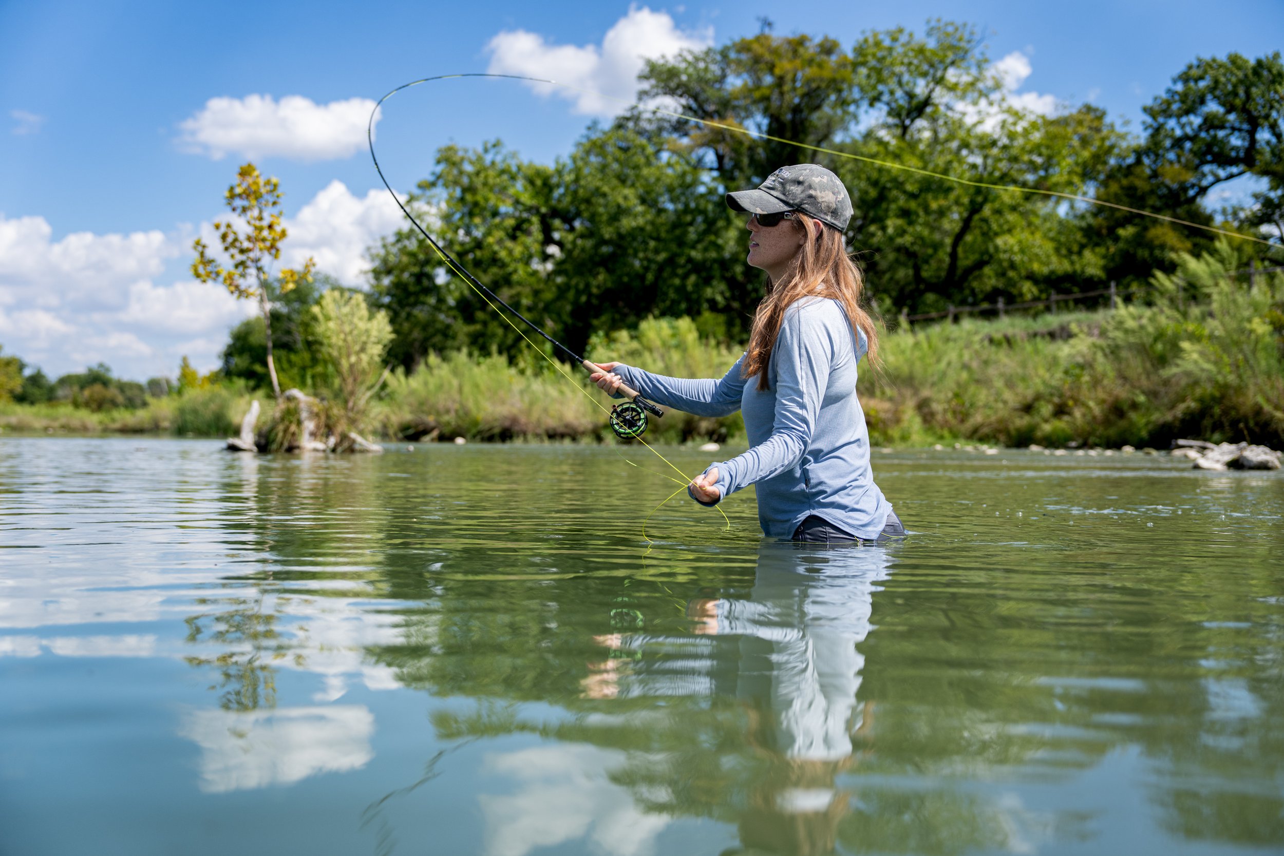 Woman in water fishing with a rod on a bright day, surrounded by trees and blue sky.