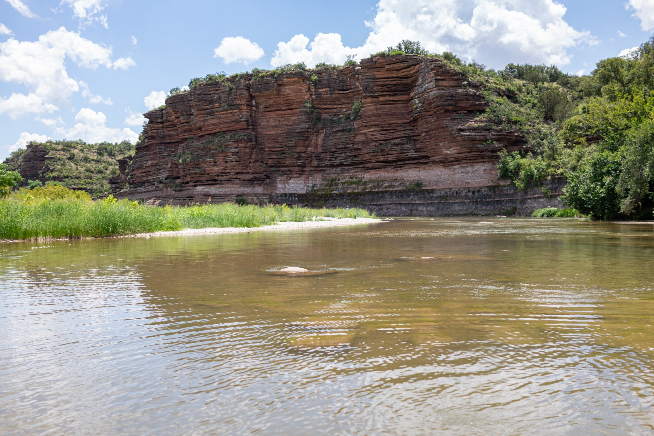 A river flowing past green vegetation with a large red rock formation in the background under a mostly cloudy sky.
