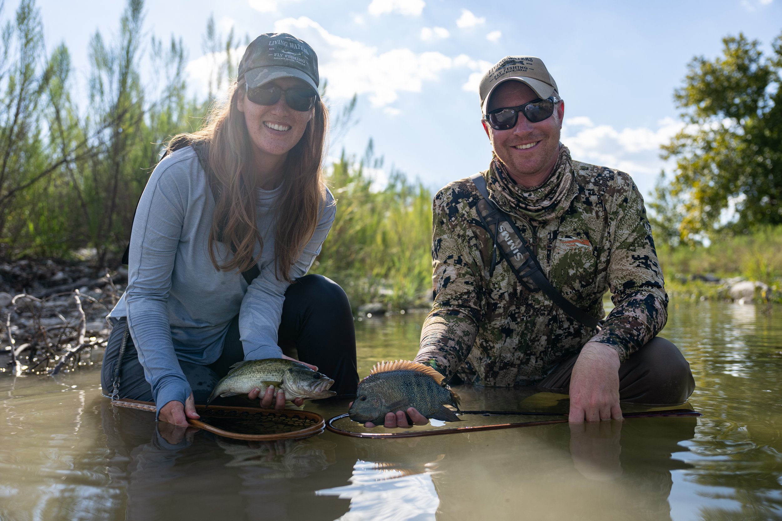 A woman and a man fishing in a shallow river, both holding fish with fishing nets, smiling at the camera on a sunny day.
