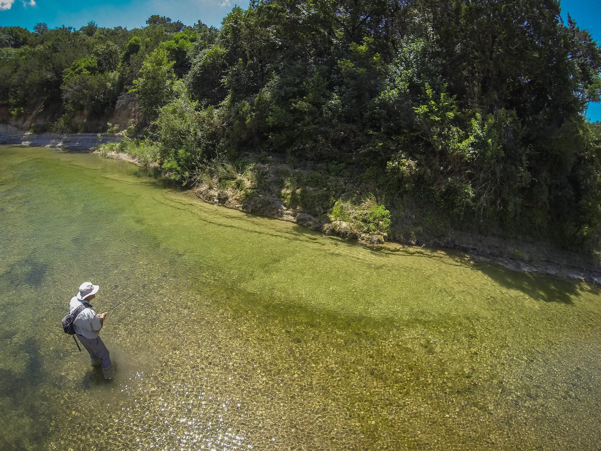 A person dressed in outdoor clothing, standing in a shallow river while fishing, with a backpack and a hat, surrounded by green trees and clear water.