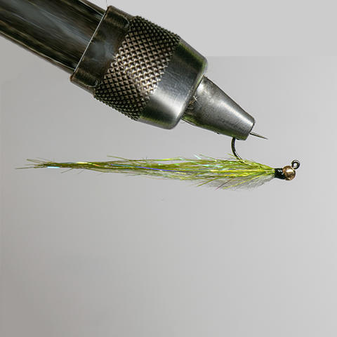 Close-up of a metal fishing lure designed to look like a small fish, attached to a drill bit, with a spinning blade and small bead at the end.