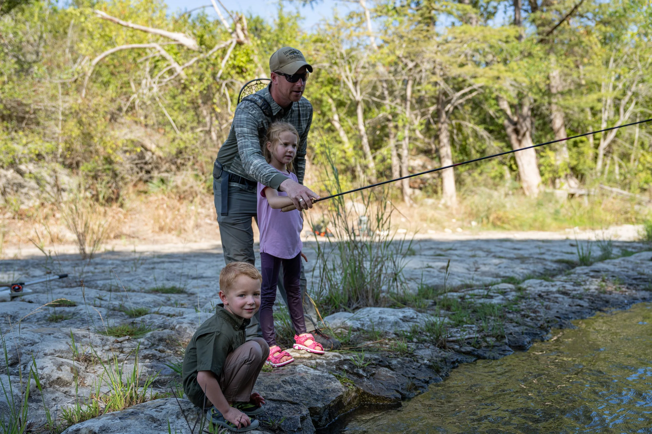 A man and two children fishing by a river in a wooded area on a sunny day.
