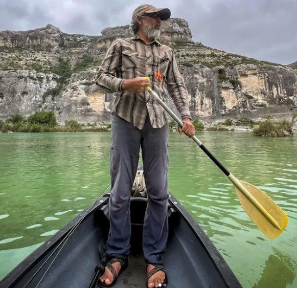 A man standing in a canoe on a calm, greenish river, holding a yellow paddle, with cliffs in the background.