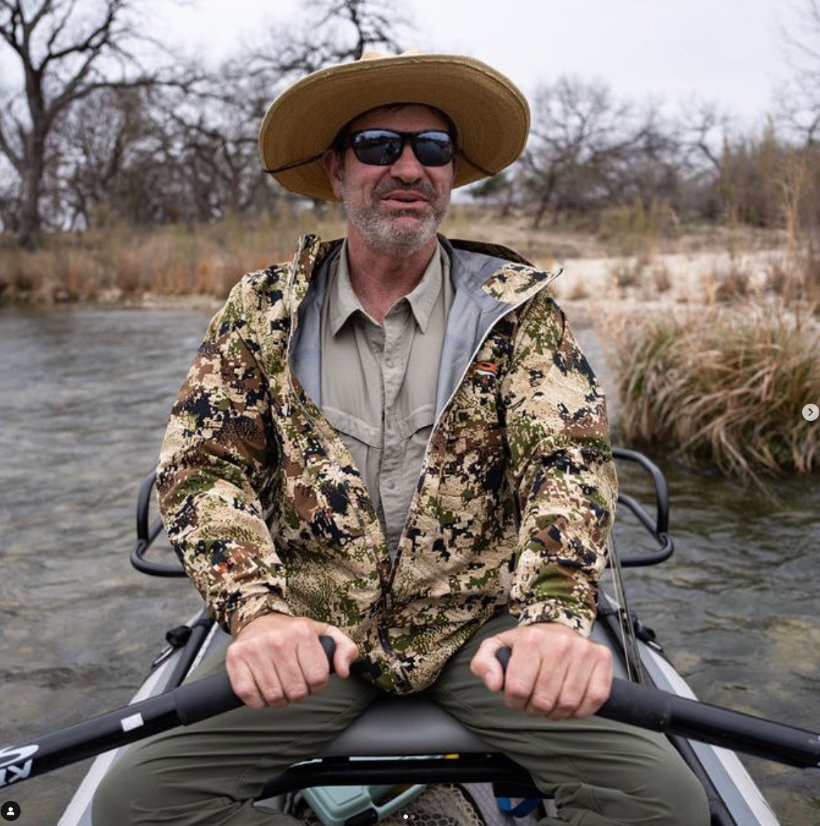 A man wearing a wide-brimmed hat, sunglasses, and a camouflage jacket, kayaking on a river with leafless trees in the background.