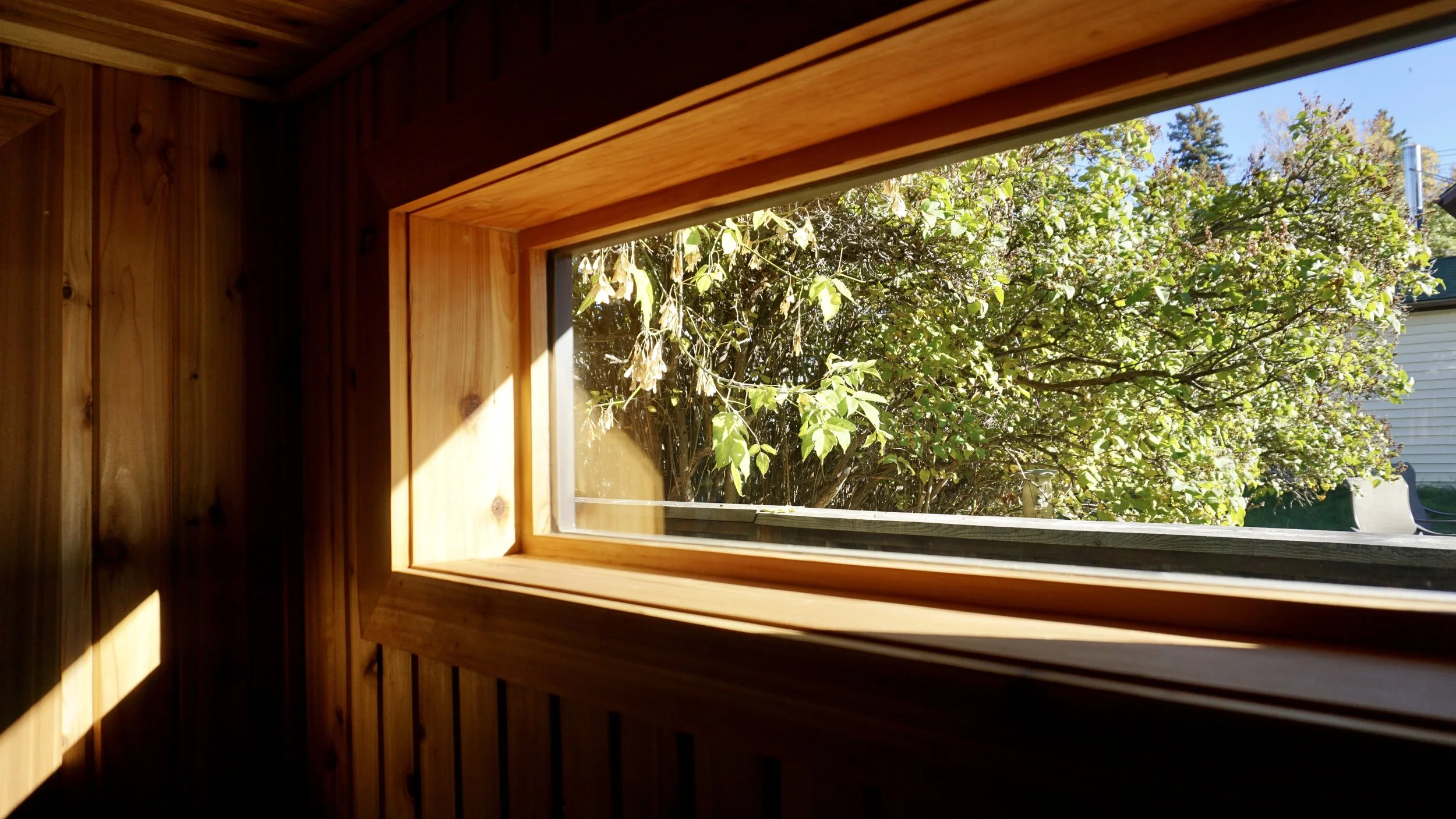 Wooden interior wall with a rectangular window, showing green trees and a white house outside on a sunny day.