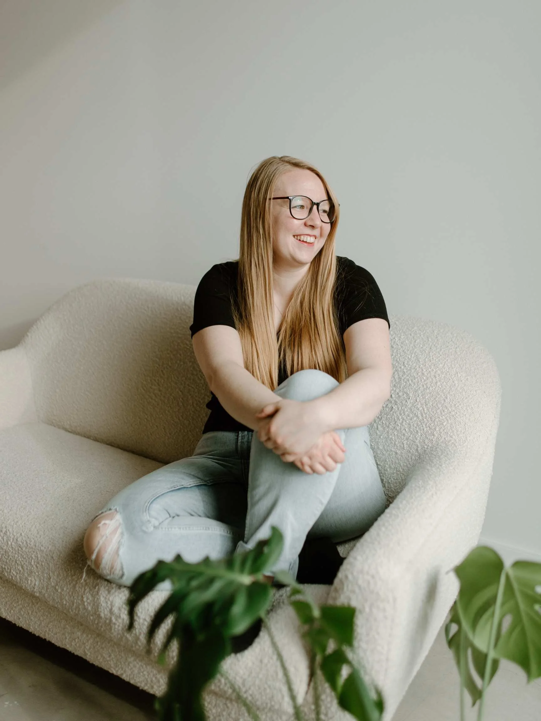 A young woman with long red hair and glasses sitting on a cream-colored sofa, smiling and looking to the side, with green houseplants in the foreground.