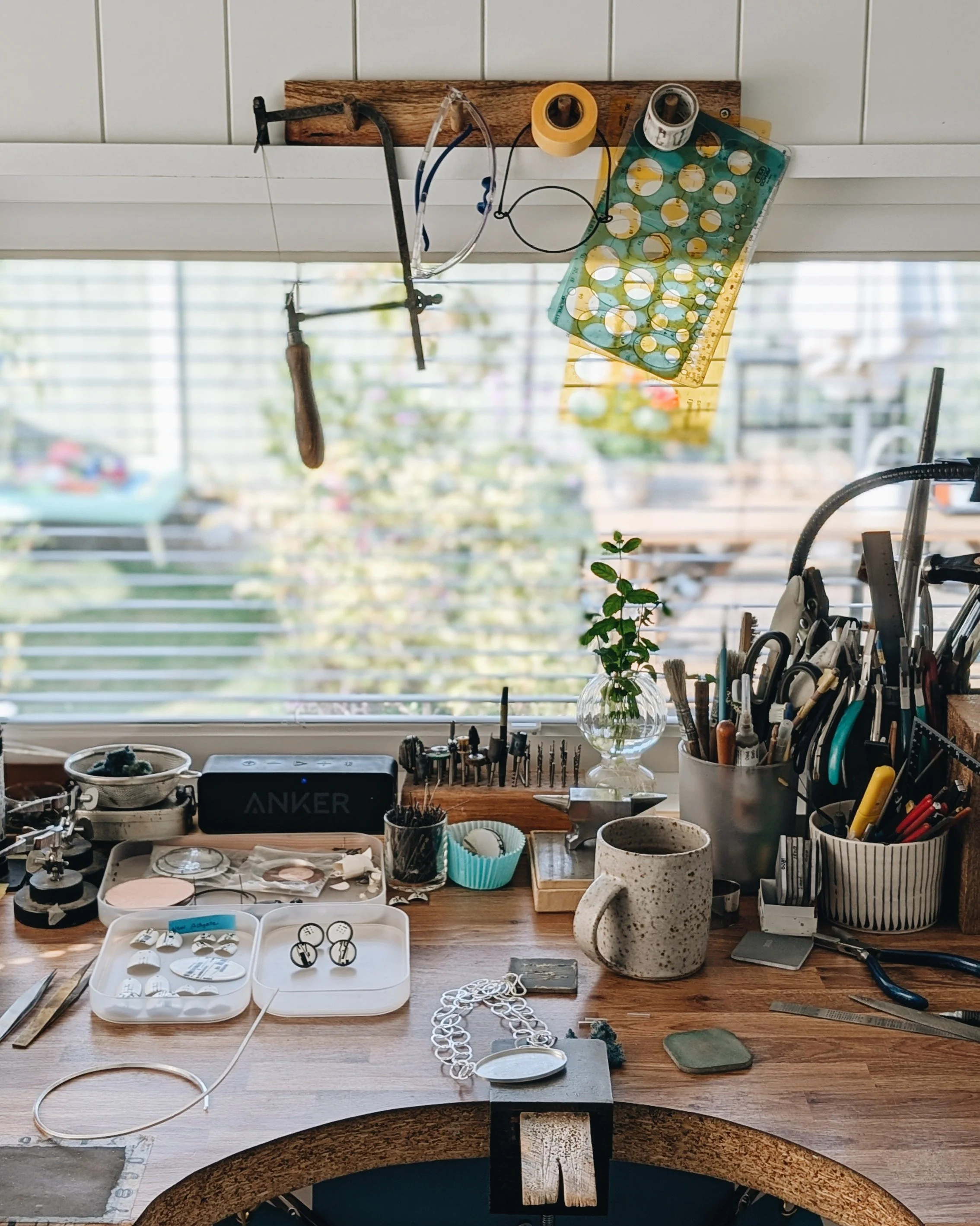 A workbench with jewelry-making tools and supplies, including a mug, jewelry pieces, pliers, and a vase with green leaves, with a window in the background.