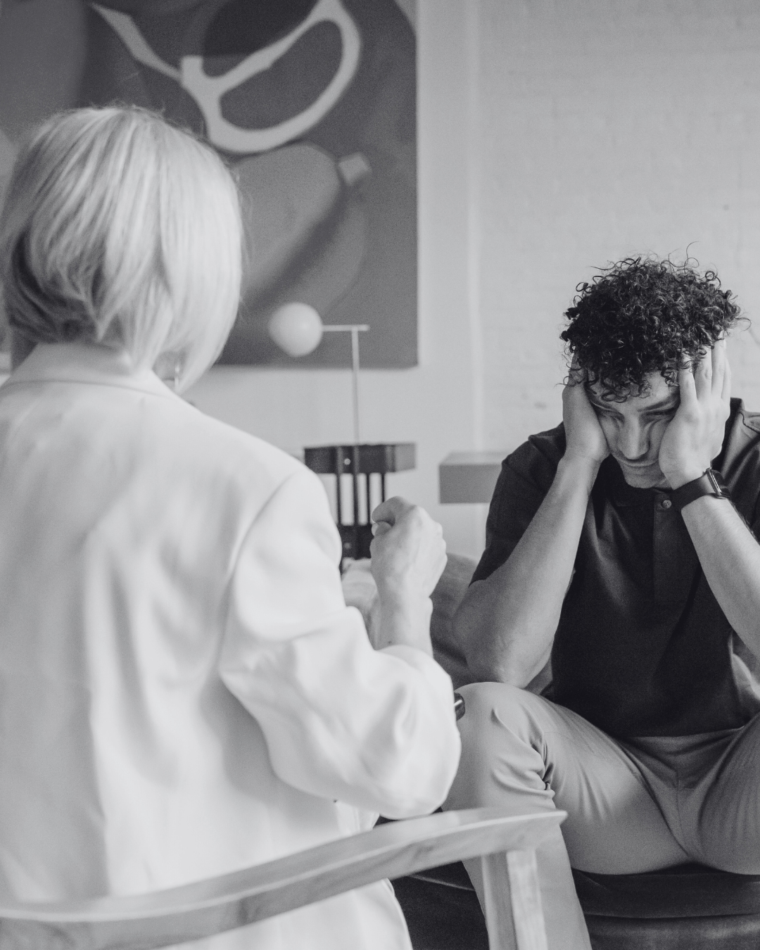 A young man with curly hair, sitting in a chair, holding his head with both hands, appears distressed. An older woman with short light-colored hair, facing away, is engaged in a conversation with him. The setting looks like a therapy or counseling session.