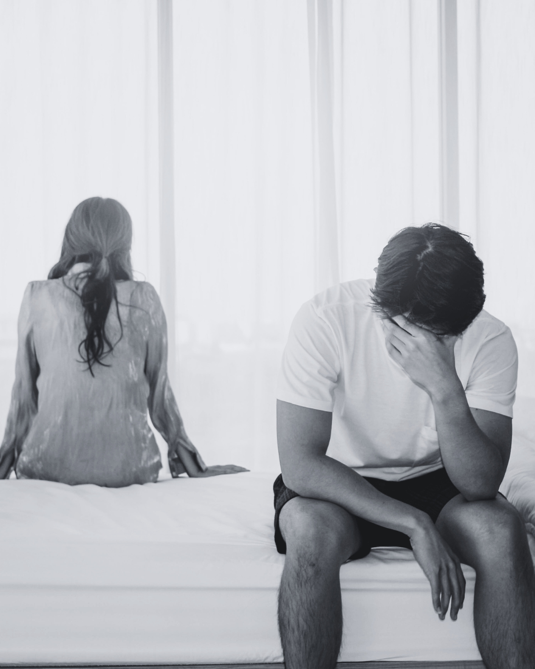 A man sitting on the bed with his head in his hand looks distressed, while a woman sits on the bed facing away from him in a bedroom with white curtains.