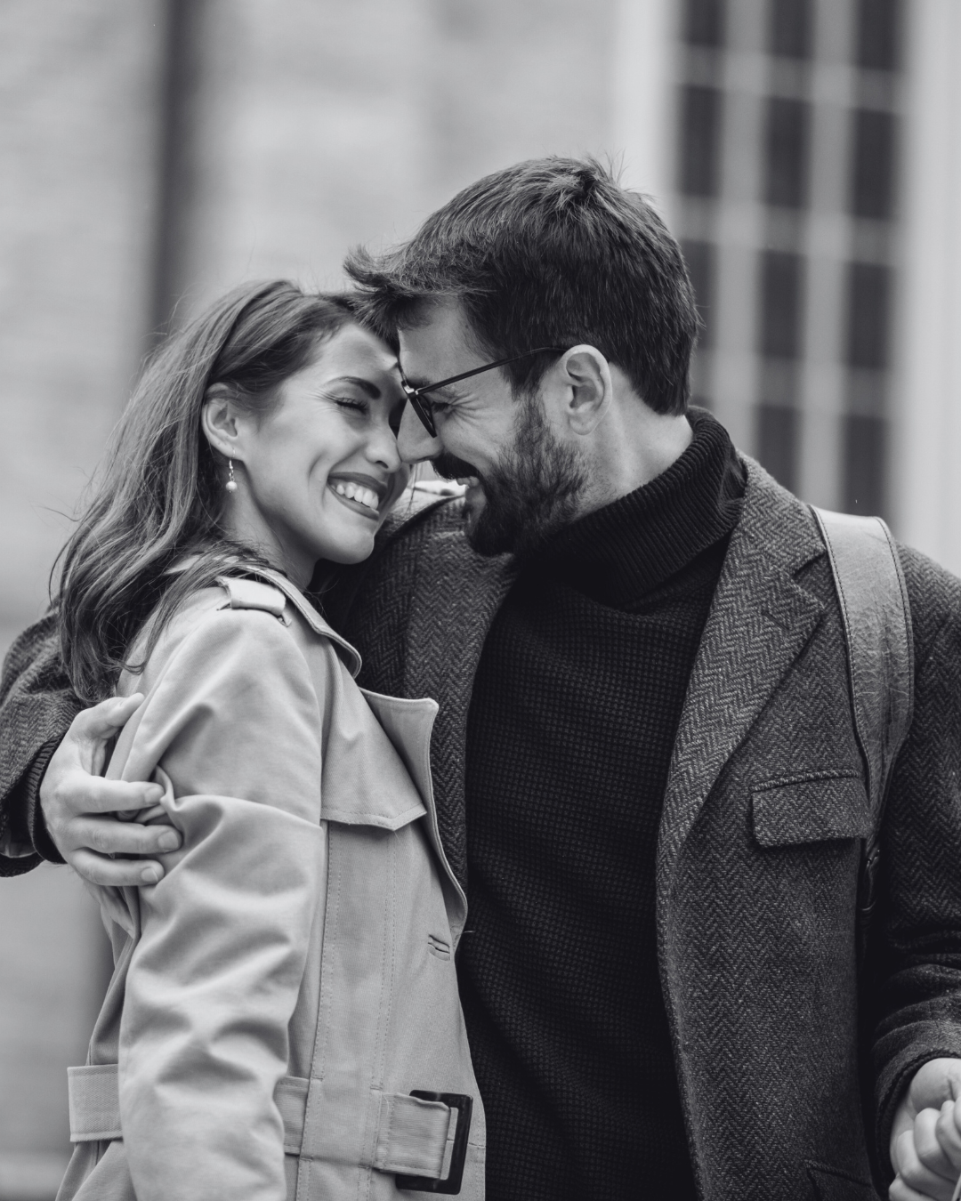 A black and white photo of a smiling couple touching foreheads and embracing outdoors.
