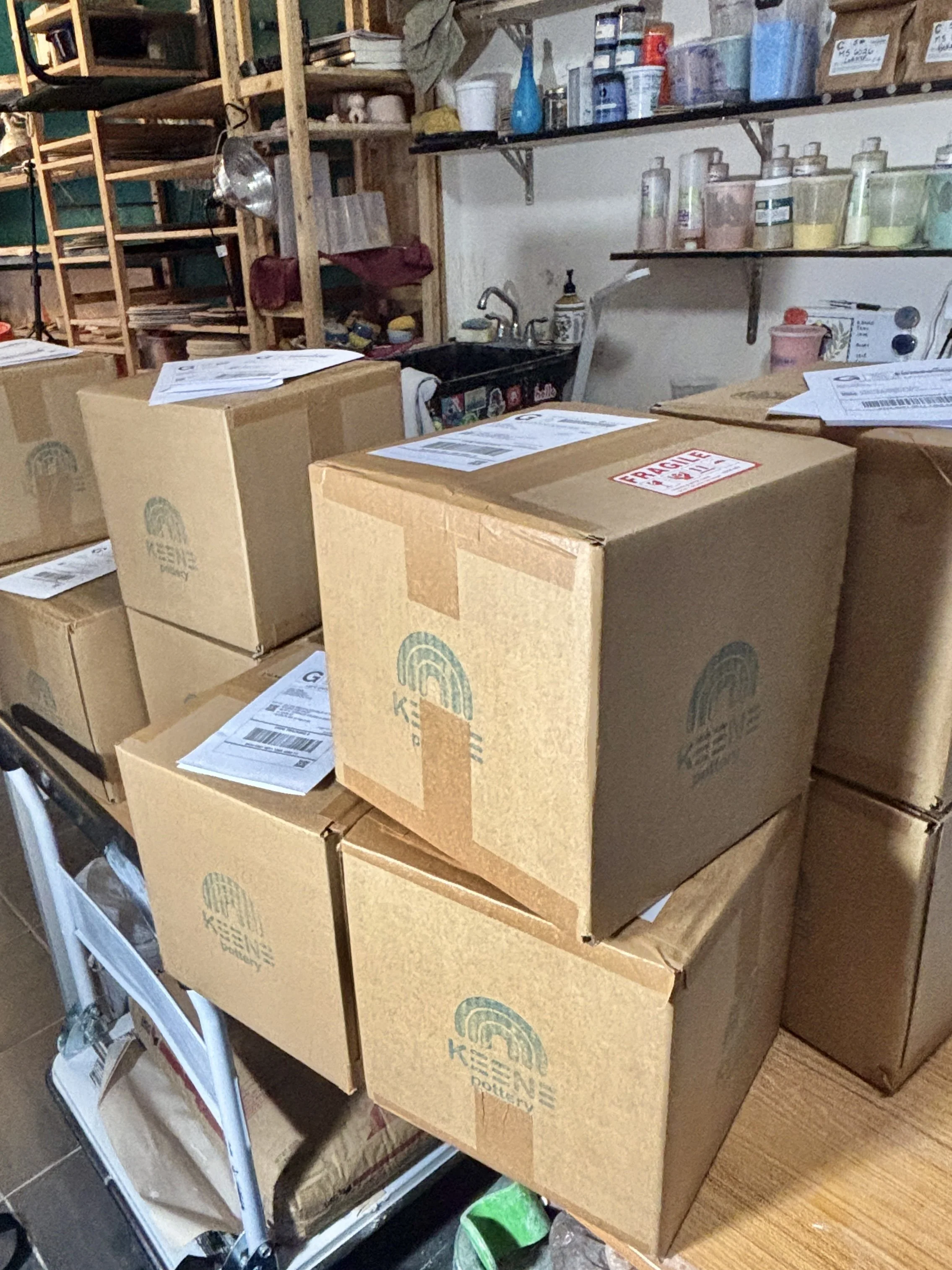 Multiple cardboard boxes with shipping labels and a rainbows logo, stacked on a cart in a storage or workshop space.