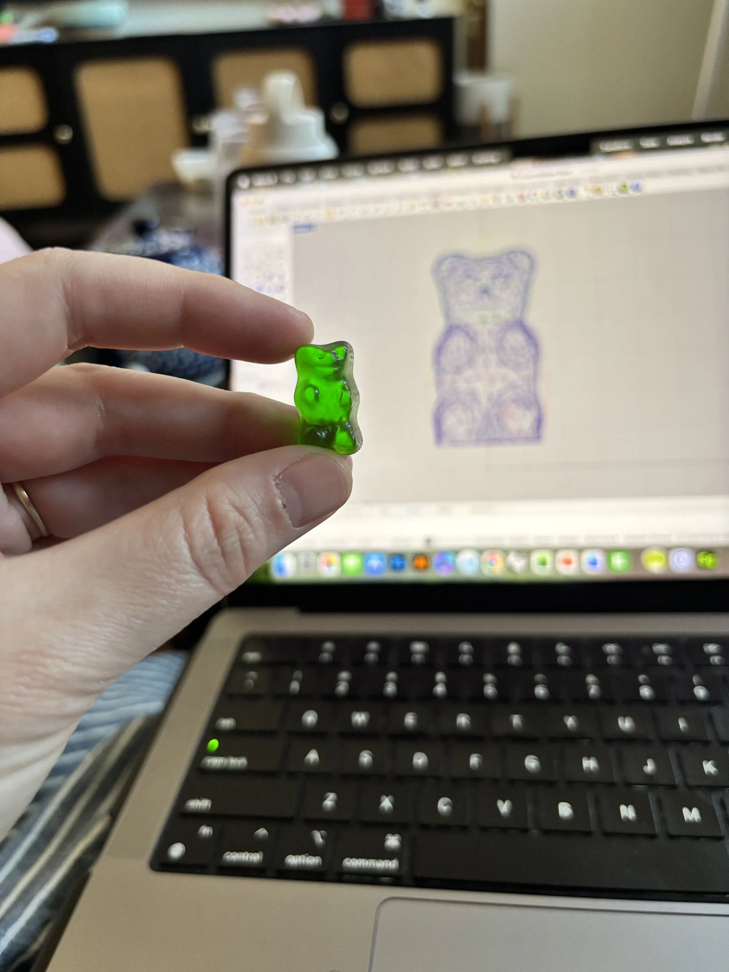 A hand holding a small green gummy bear in front of a laptop with an open drawing program, displaying a purple outline of the gummy bear on the screen, in a room with a wooden cabinet and some items in the background.