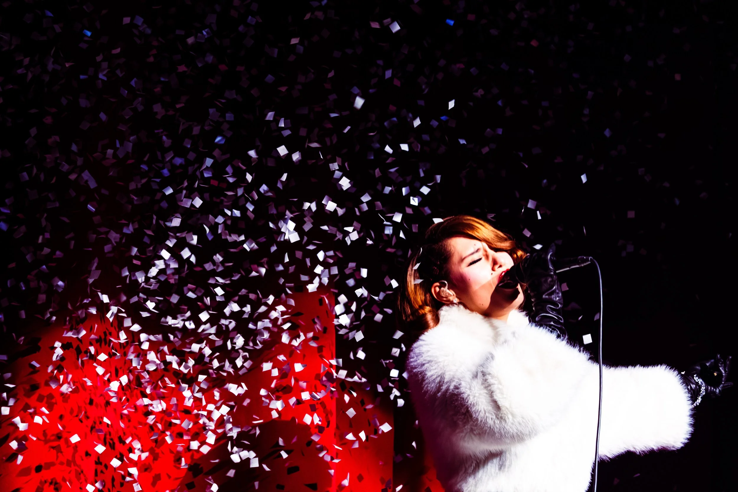 A woman singing into a microphone on stage with black and white confetti falling around her, wearing a white fur coat and black gloves.