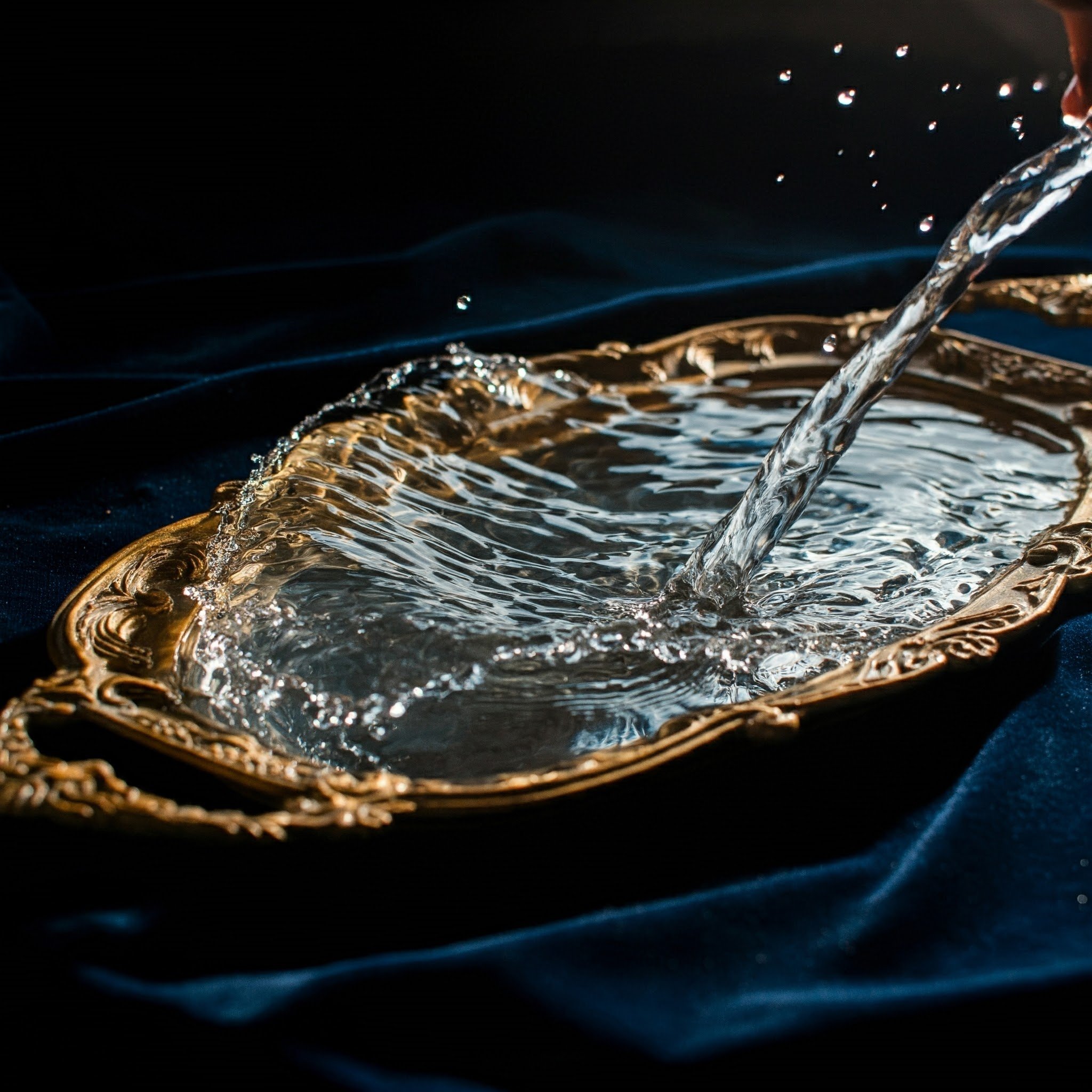 Stream of water pouring into a gold tray on a dark blue velvet cloth, symbolizing the refreshing and luxurious essence of the Aqua Wave Grasse scent