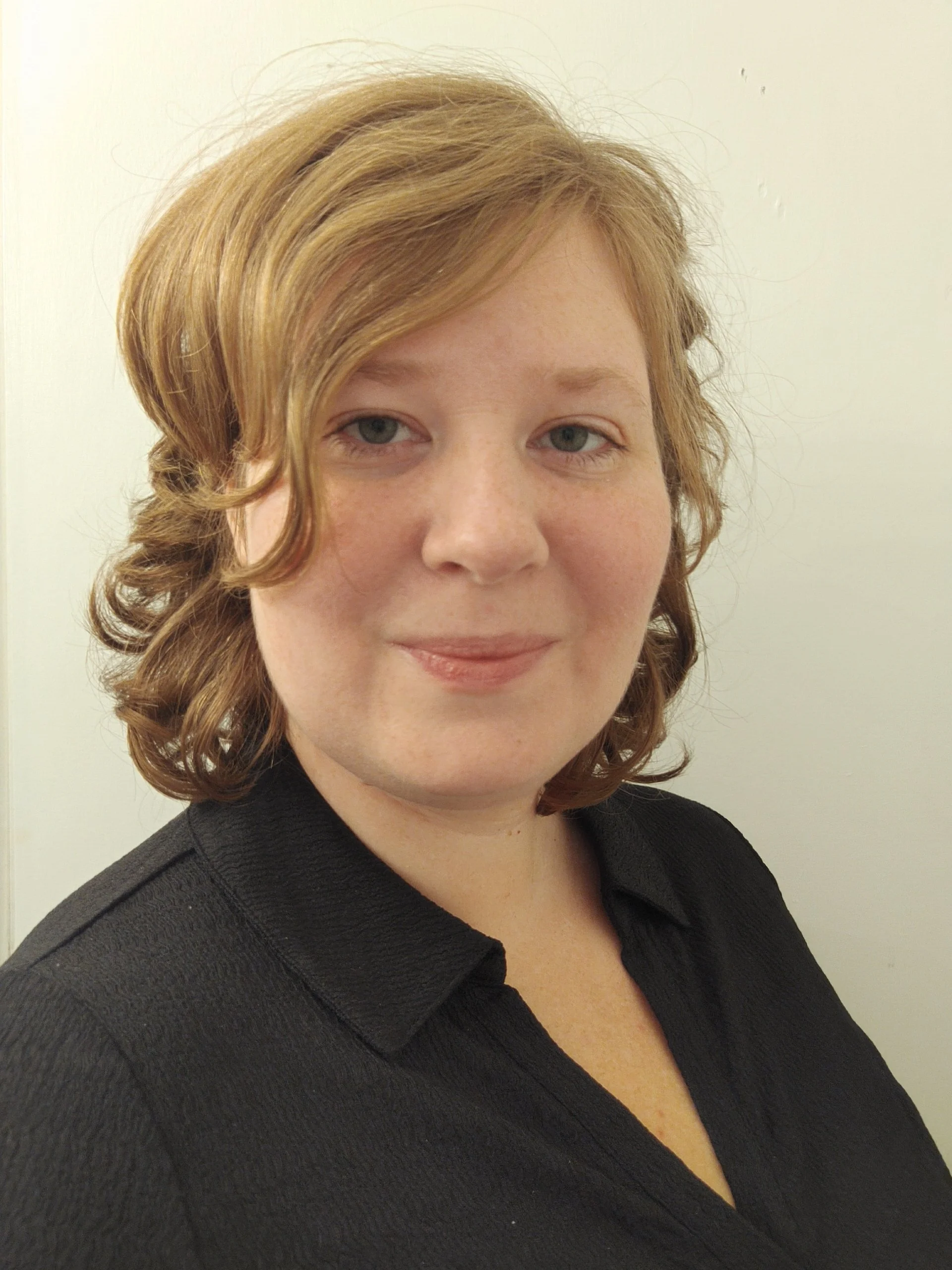 Close-up of a woman with wavy light brown hair and fair skin, wearing a black top, standing against a plain white wall.