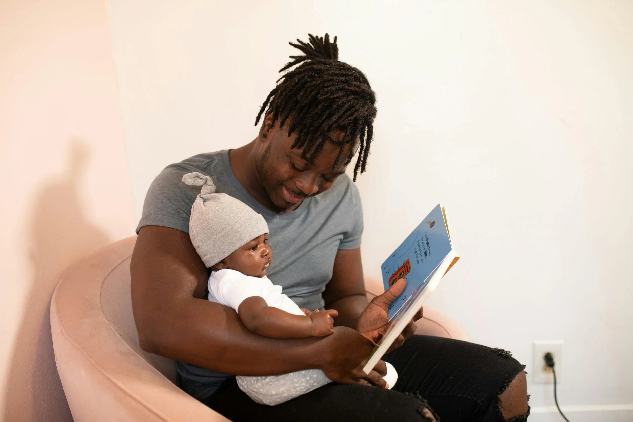 A man with dreadlocks, wearing a gray T-shirt and ripped black jeans, sitting on a pink chair, holding a baby on his lap while reading a book. The baby is wearing a gray hat and white outfit, looking at the book.