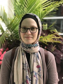 Woman wearing glasses and a patterned scarf standing in front of tropical plants.