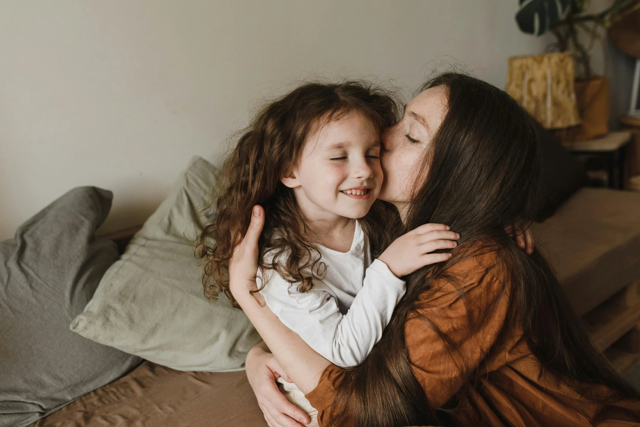 A woman with long dark hair and a young girl with curly red hair hugging and kissing each other in a cozy living room.