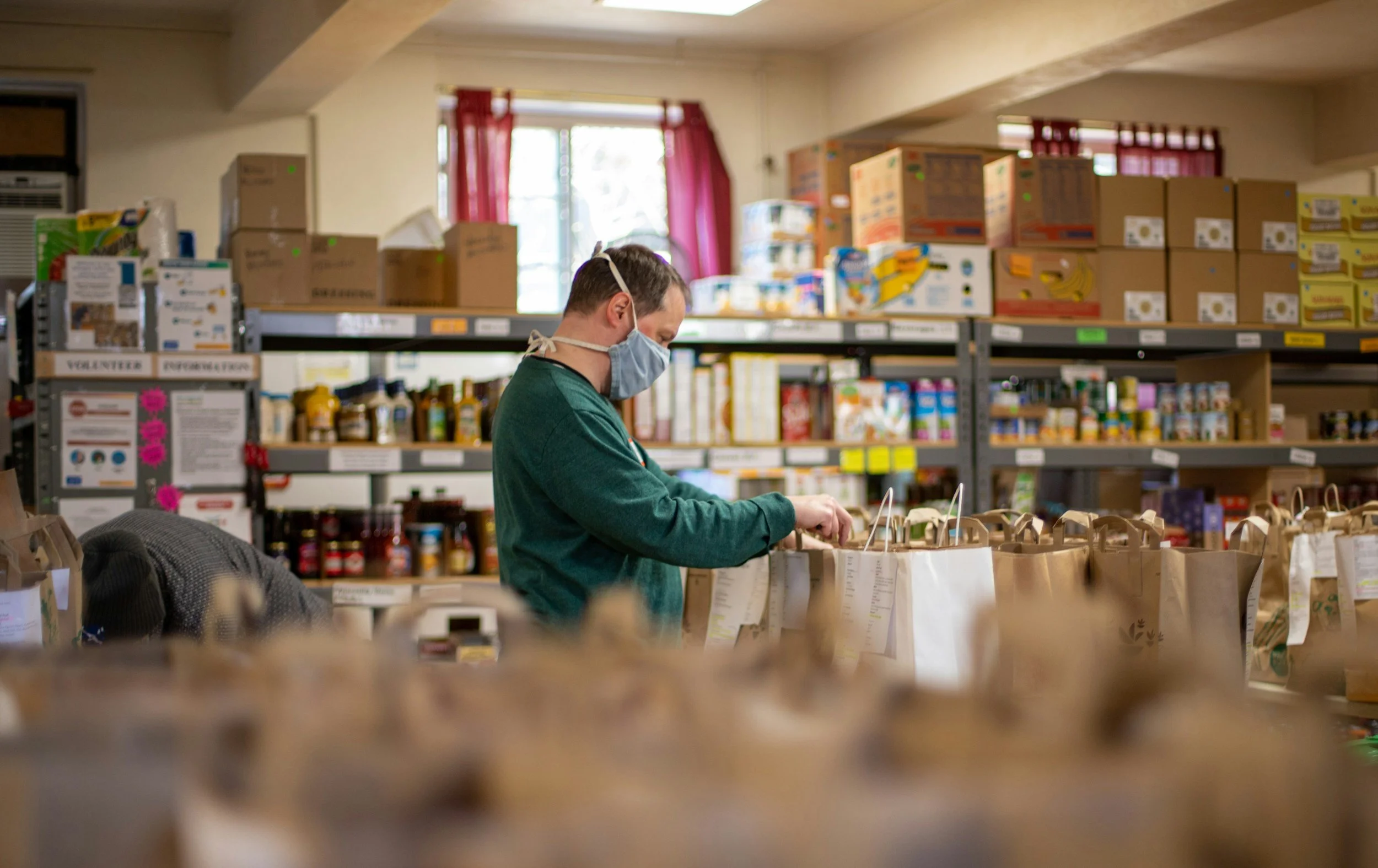 A person wearing a face mask and gloves shopping in a grocery store, browsing through paper bags filled with products, with shelves stocked with various food items and boxes in the background.