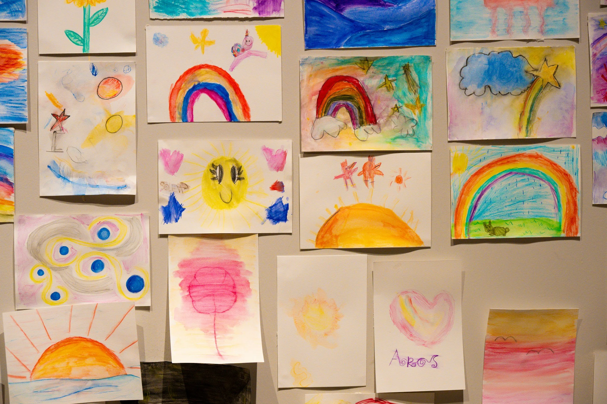 Collection of children's colorful drawings on paper, featuring rainbows, sun, hearts, and abstract shapes.