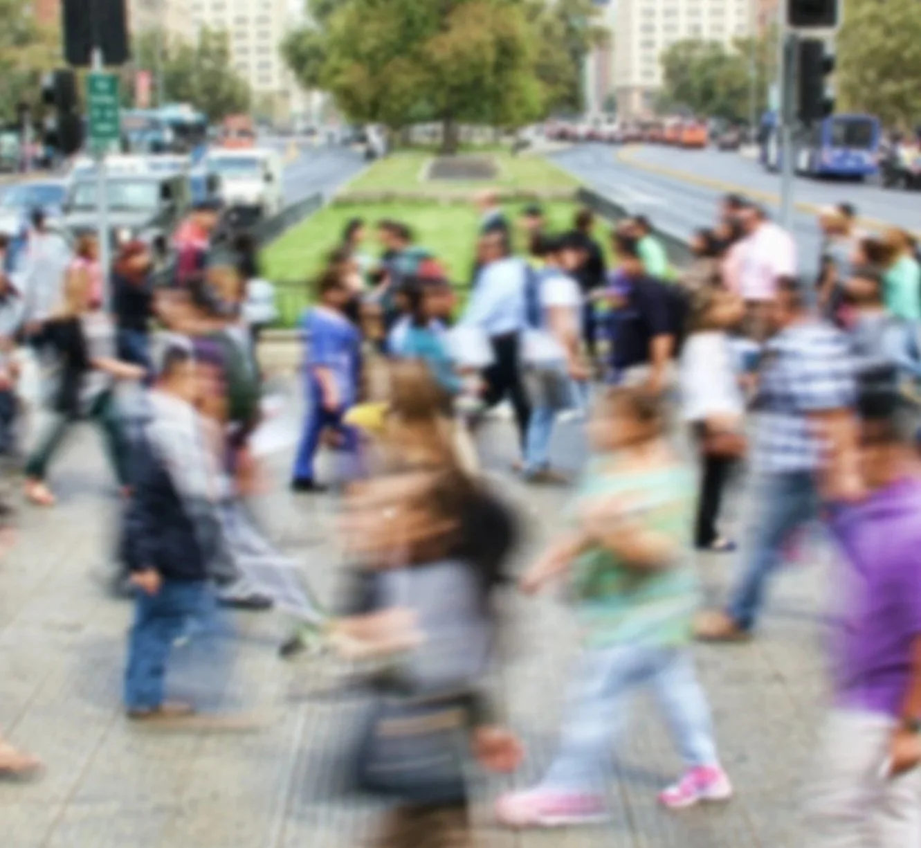 A busy city intersection with people walking in multiple directions, trees, and buildings in the background.