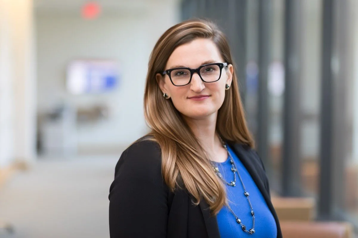 A woman with long brown hair, wearing glasses, a blue top, a black blazer, and jewelry, standing indoors with a blurred background.