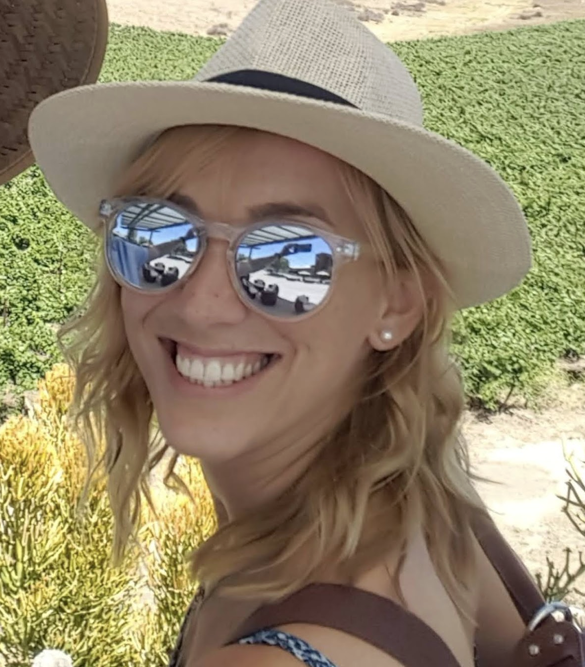 A woman with blonde wavy hair smiling outdoors, wearing a white sun hat, mirrored sunglasses, and pearl earrings, with greenery and a sunny sky reflected in her sunglasses.