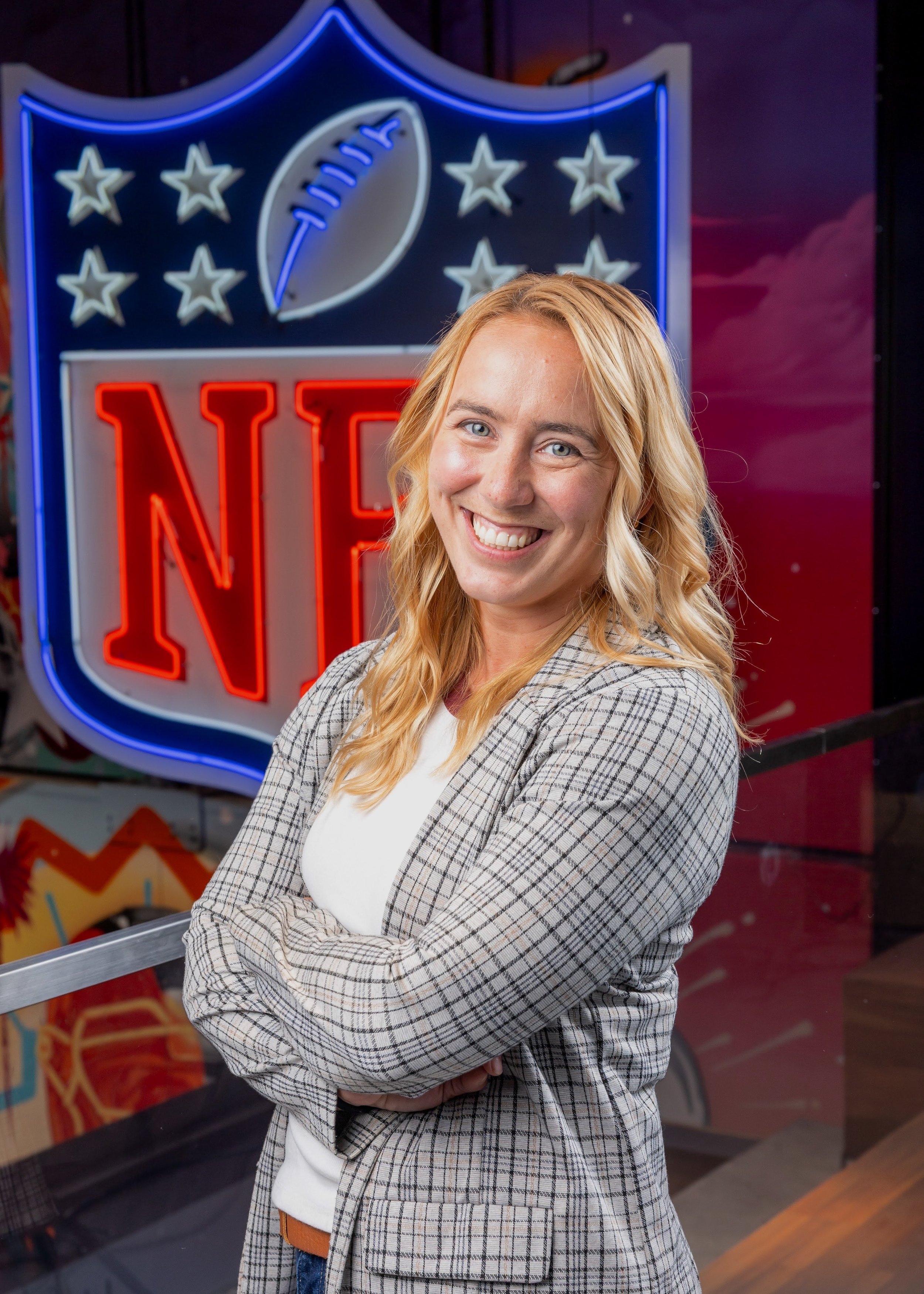 A smiling woman with blonde hair wearing a plaid blazer standing in front of an illuminated NFL logo sign.