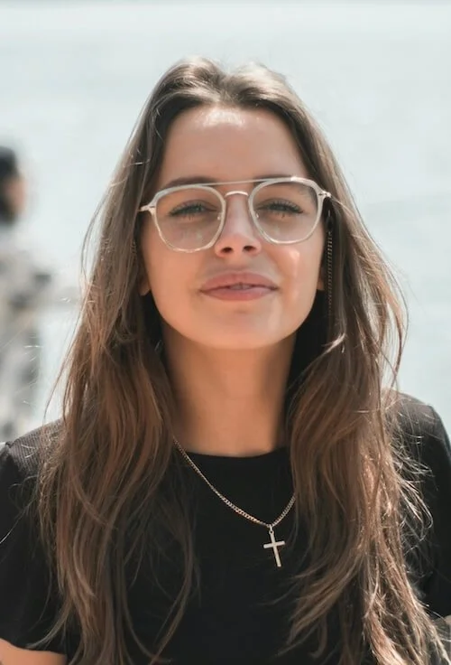 Close-up of a young woman with long brown hair wearing glasses, a necklace with a cross pendant, and a black top, outdoors near water.