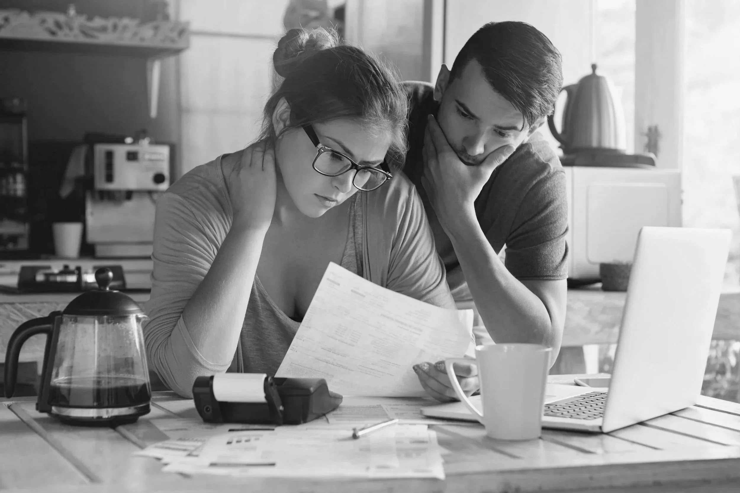Financial wellness coaching for A distressed woman with glasses and a man appear frustrated while reviewing paperwork at a kitchen table with a laptop, coffee mug, and a calculator.